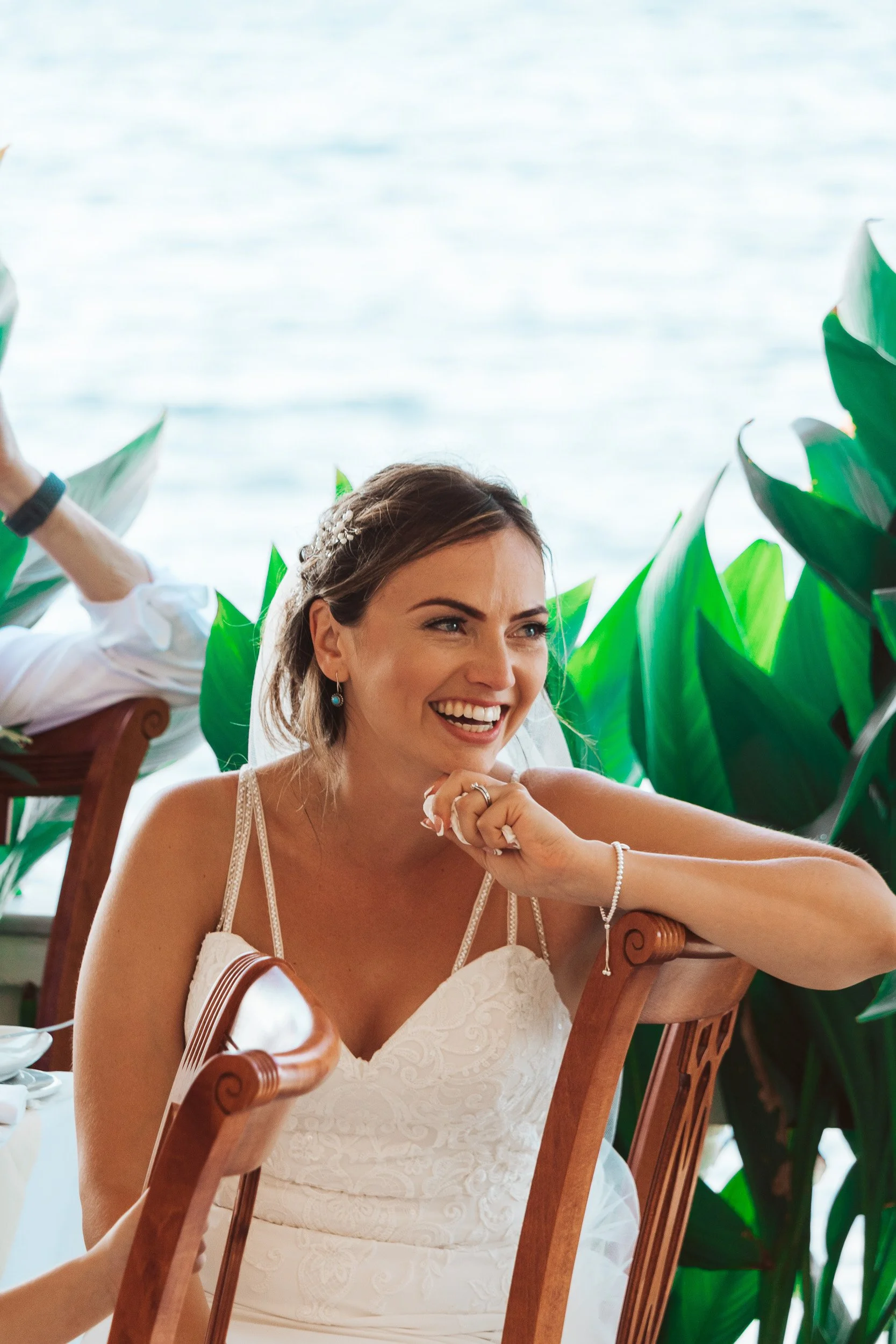 Smiling bride in a white wedding dress sitting at a wooden table with green plants and water in the background.