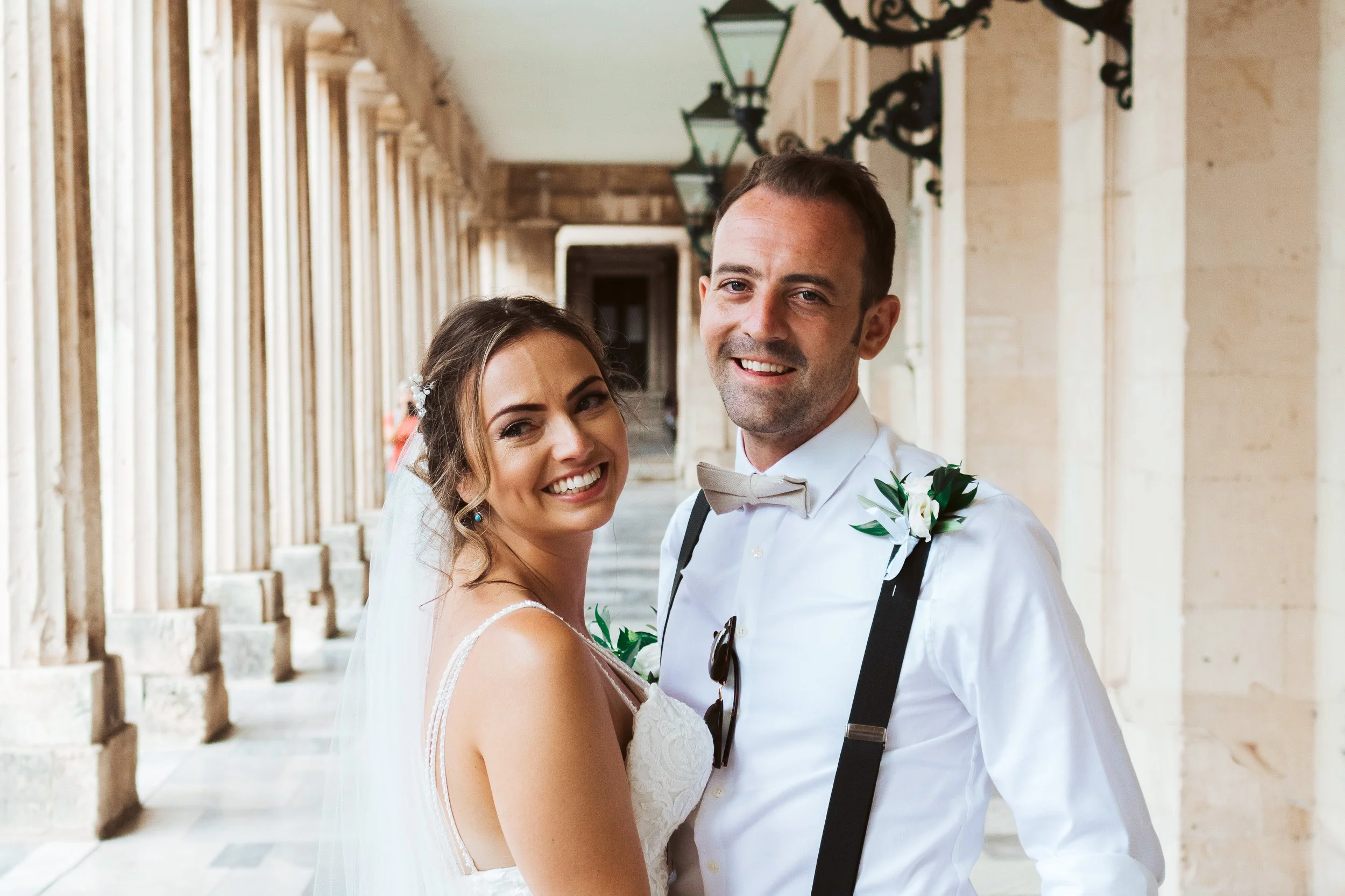 A smiling bride and groom standing together in an outdoor corridor, with columns and hanging lanterns.