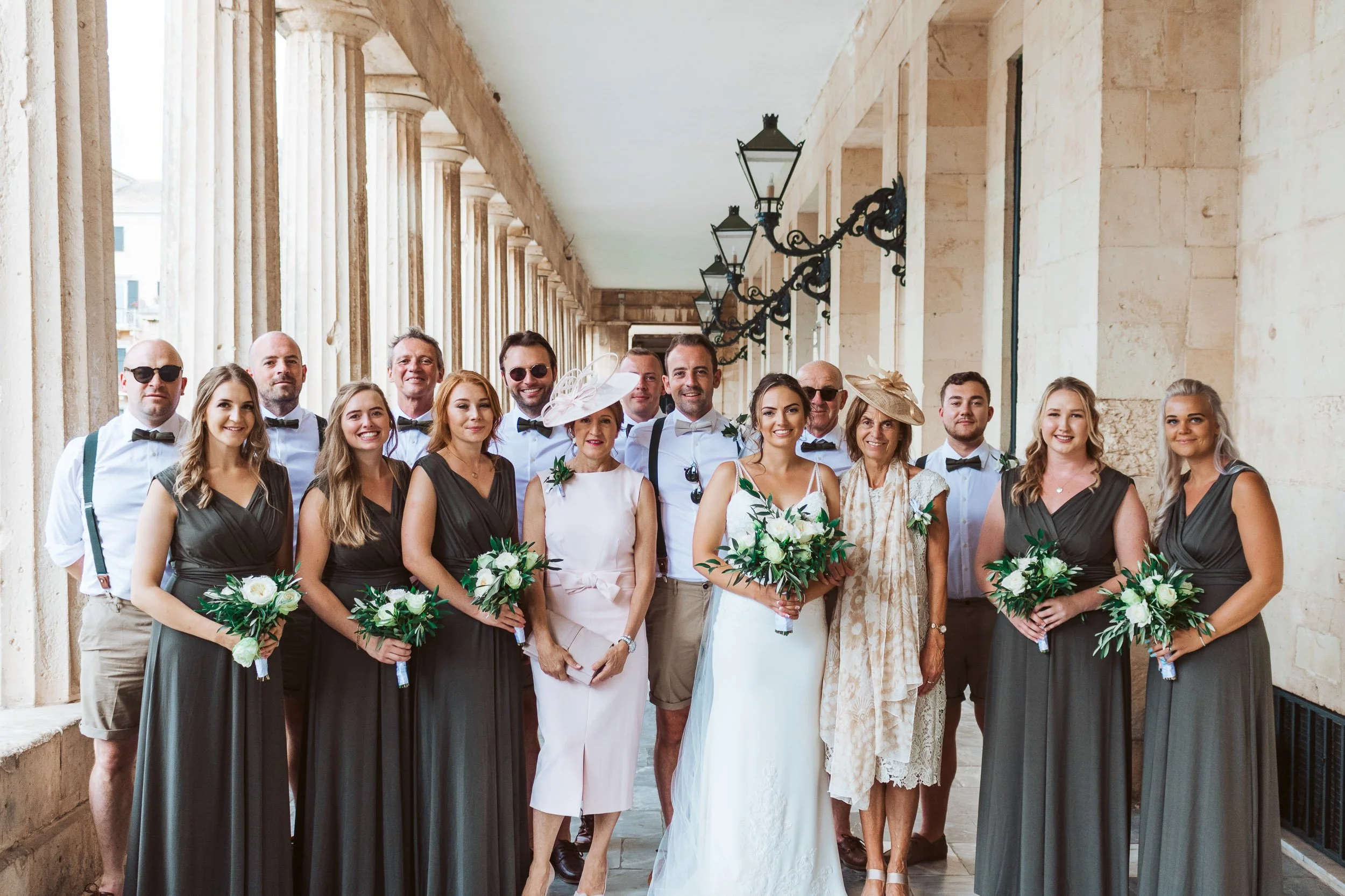 Group of wedding guests and bridal party standing under an arched walkway with stone walls, some holding bouquets, smiling at the camera.