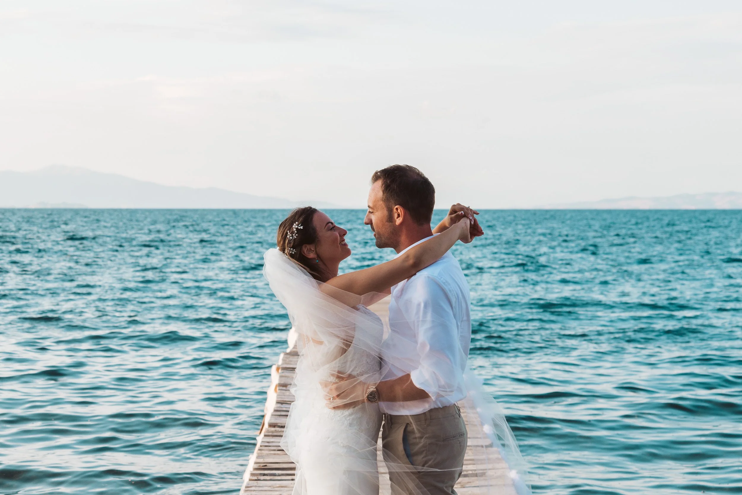 A bride and groom stand on a wooden dock by the water, embracing and smiling at each other, with a distant mountain visible across the water.