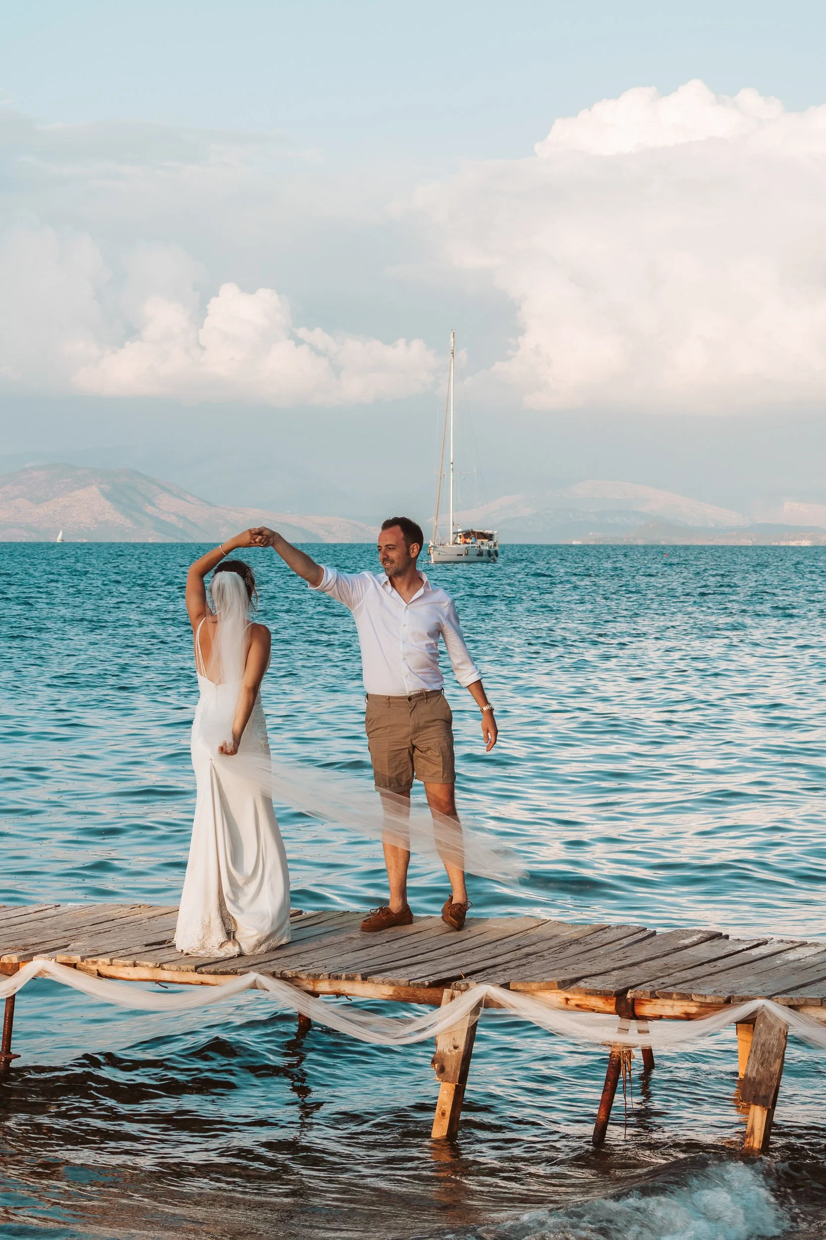 A couple dancing on a wooden dock by the sea with a sailboat in the background and mountains on the horizon.