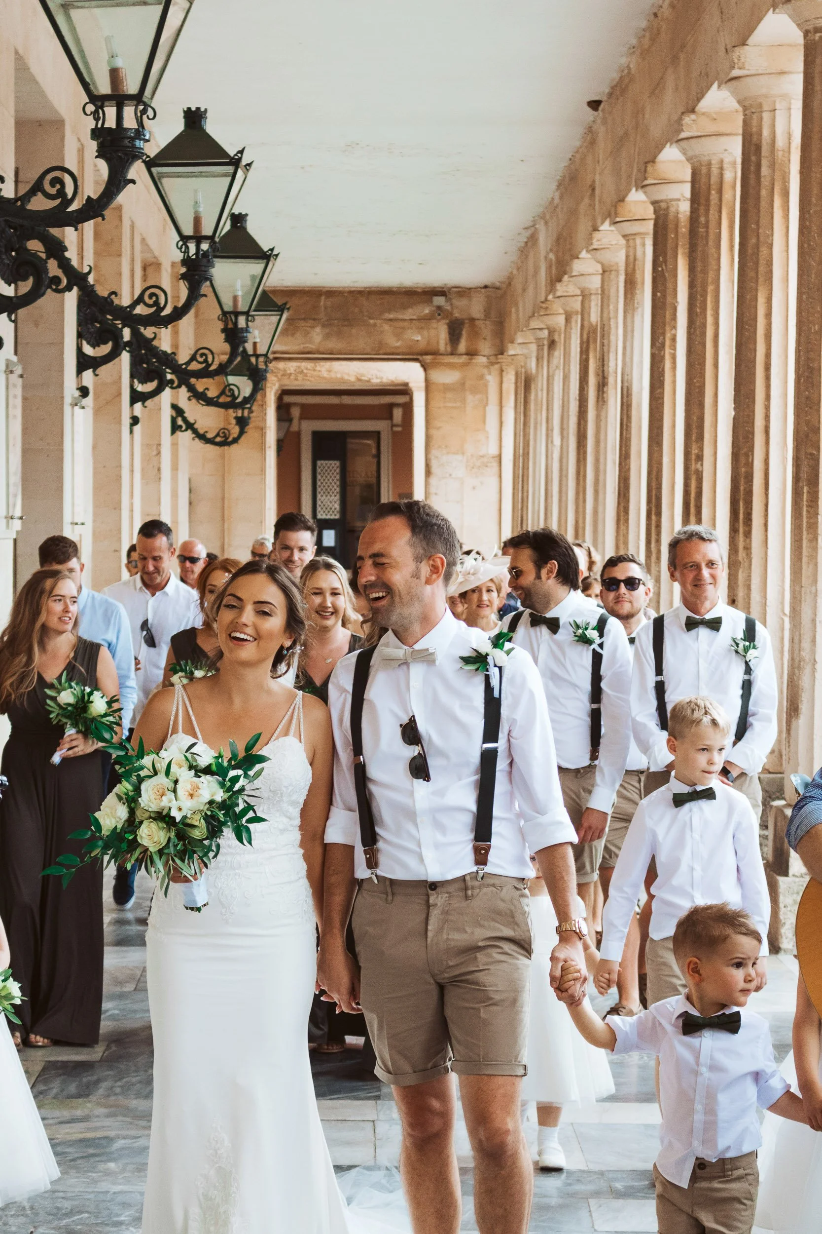 A wedding procession with the bride and groom leading, surrounded by family and friends, walking through a stone corridor with vintage light fixtures.