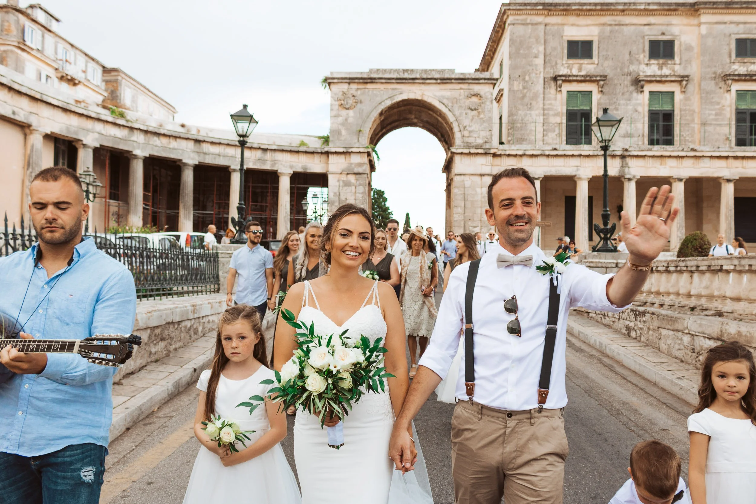 A group of people, including a bride and groom, walking outdoors during a wedding celebration. The bride is holding a bouquet of white roses, and the groom is waving. There are children and other guests in the background, with historic buildings and 