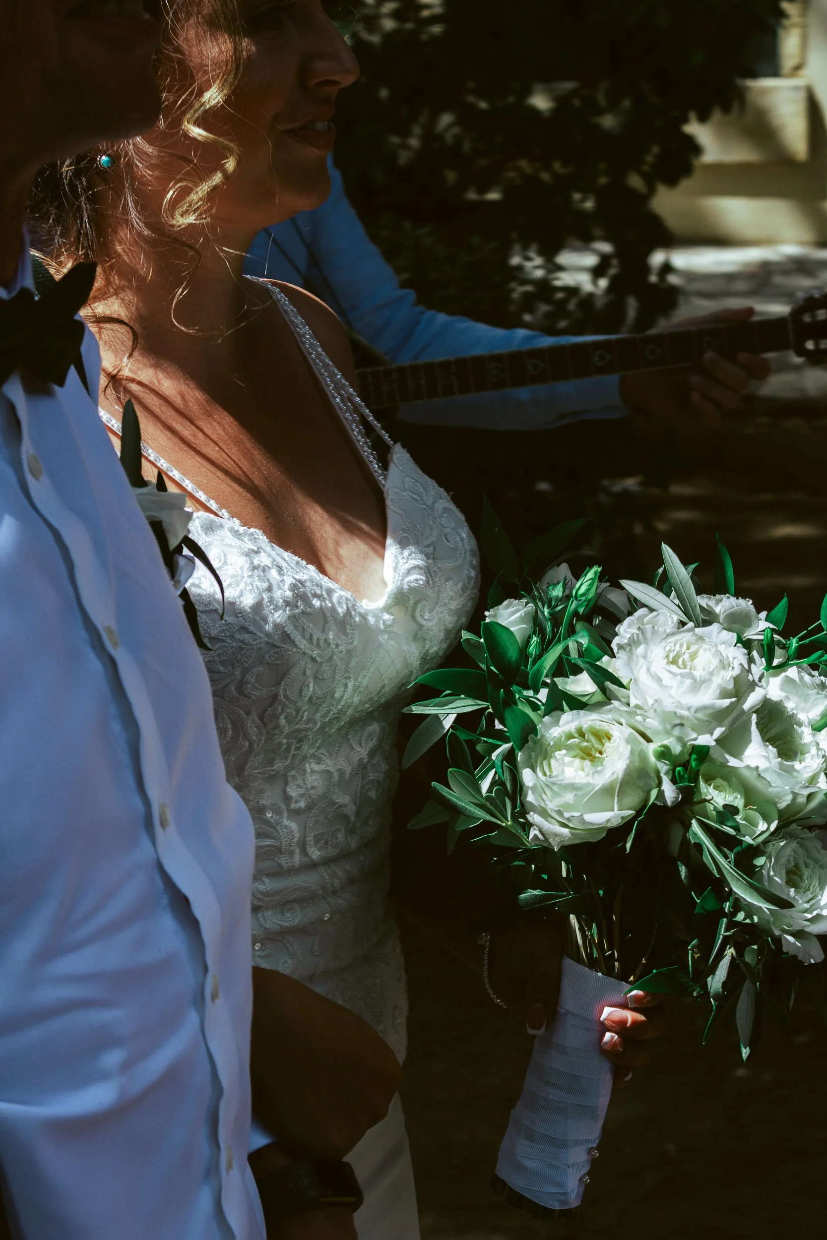 A bride holding a bouquet of white roses and greenery, with a woman beside her and a musician playing guitar in the background.