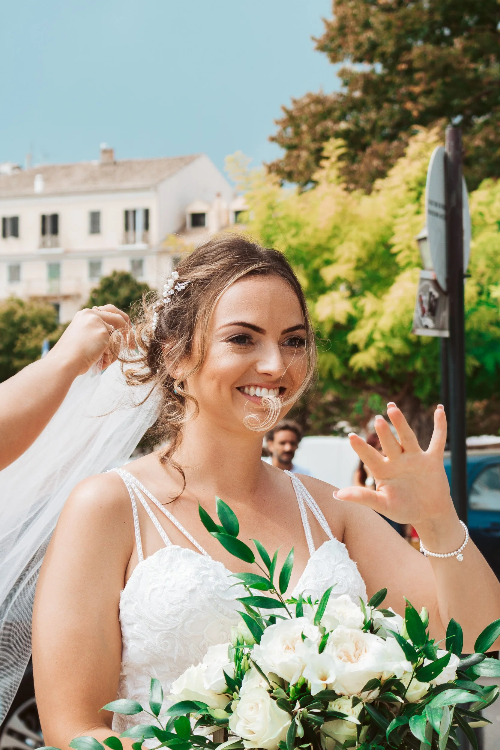 Bride smiling and waving in a wedding dress holding a bouquet of white roses with greenery outdoors, with trees and buildings in the background.