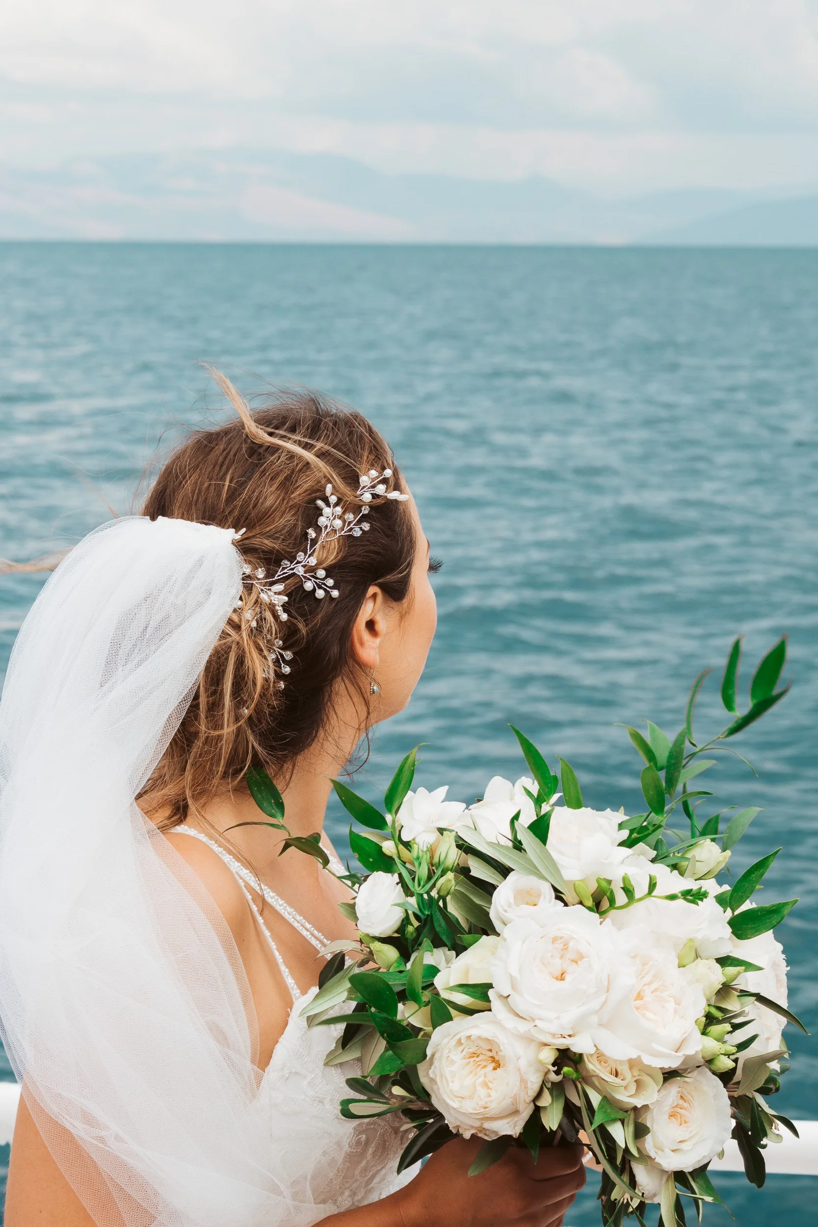 Bride holding a bouquet of white flowers on a boat, looking out at the ocean.