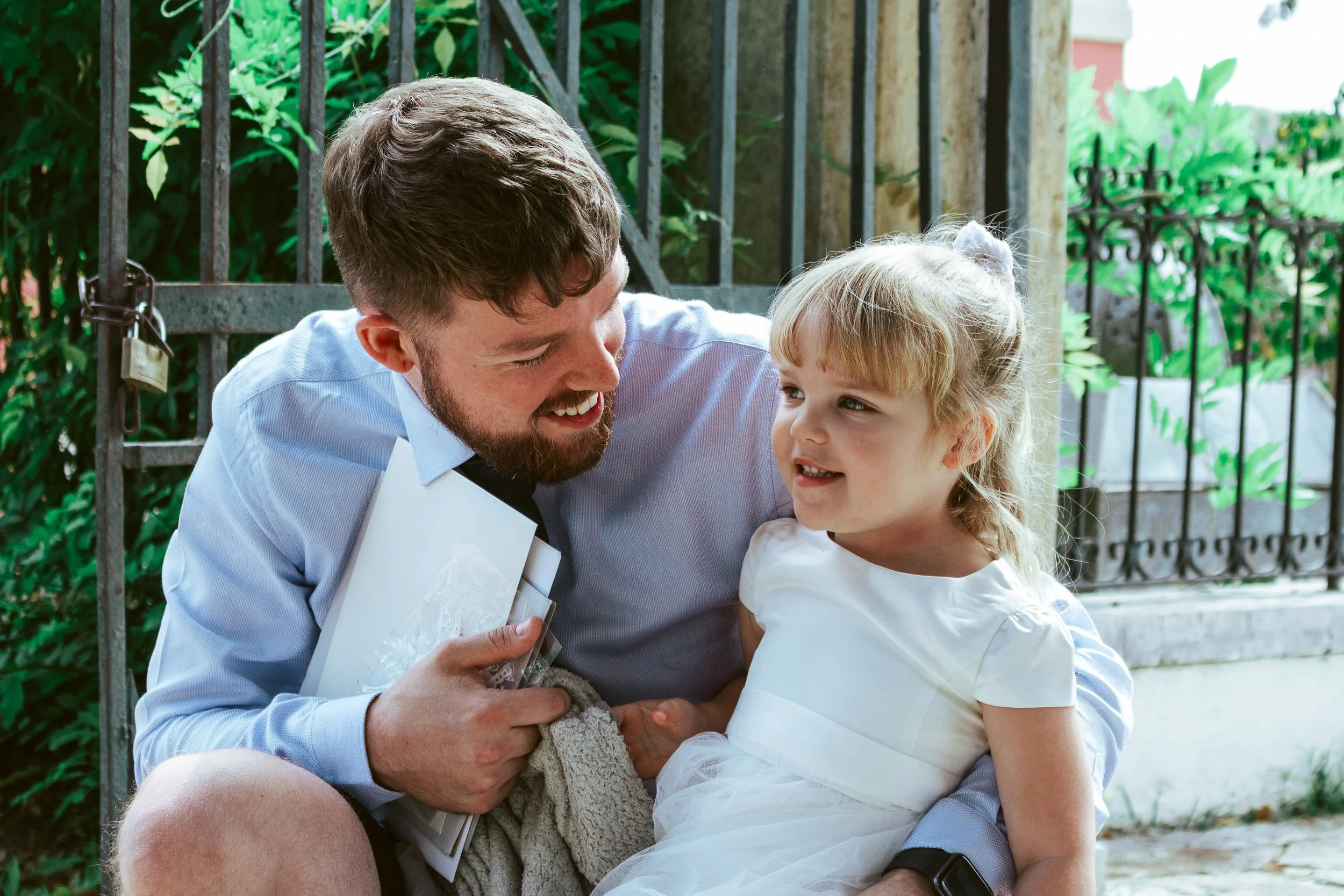 A man and young girl sharing a joyful moment outdoors, with a gated fence and greenery in the background.