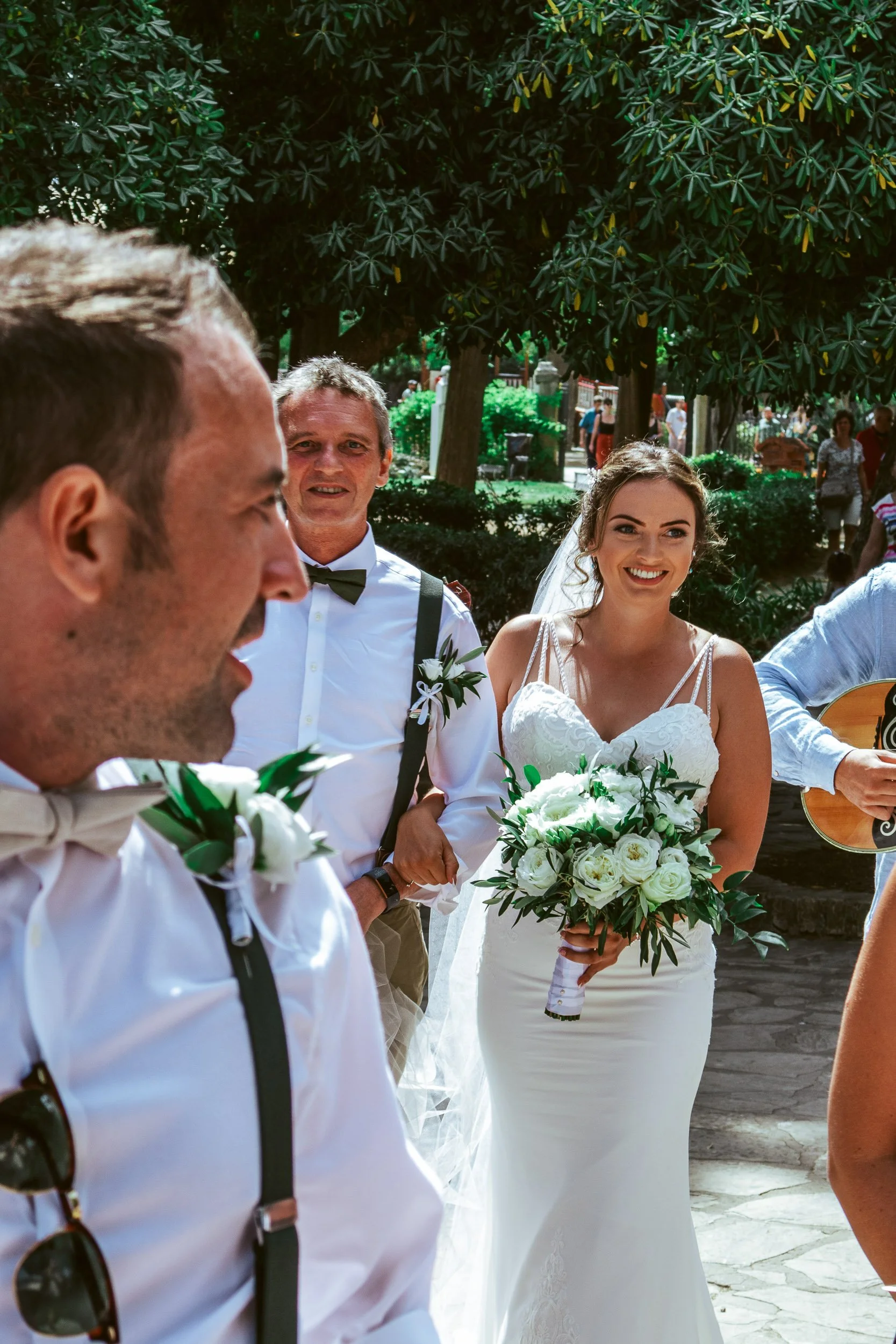 A bride in a white wedding dress holding a bouquet of white roses and greenery, smiling, with men in formal attire and sunglasses, outdoors in a sunny park with trees and other people in the background.
