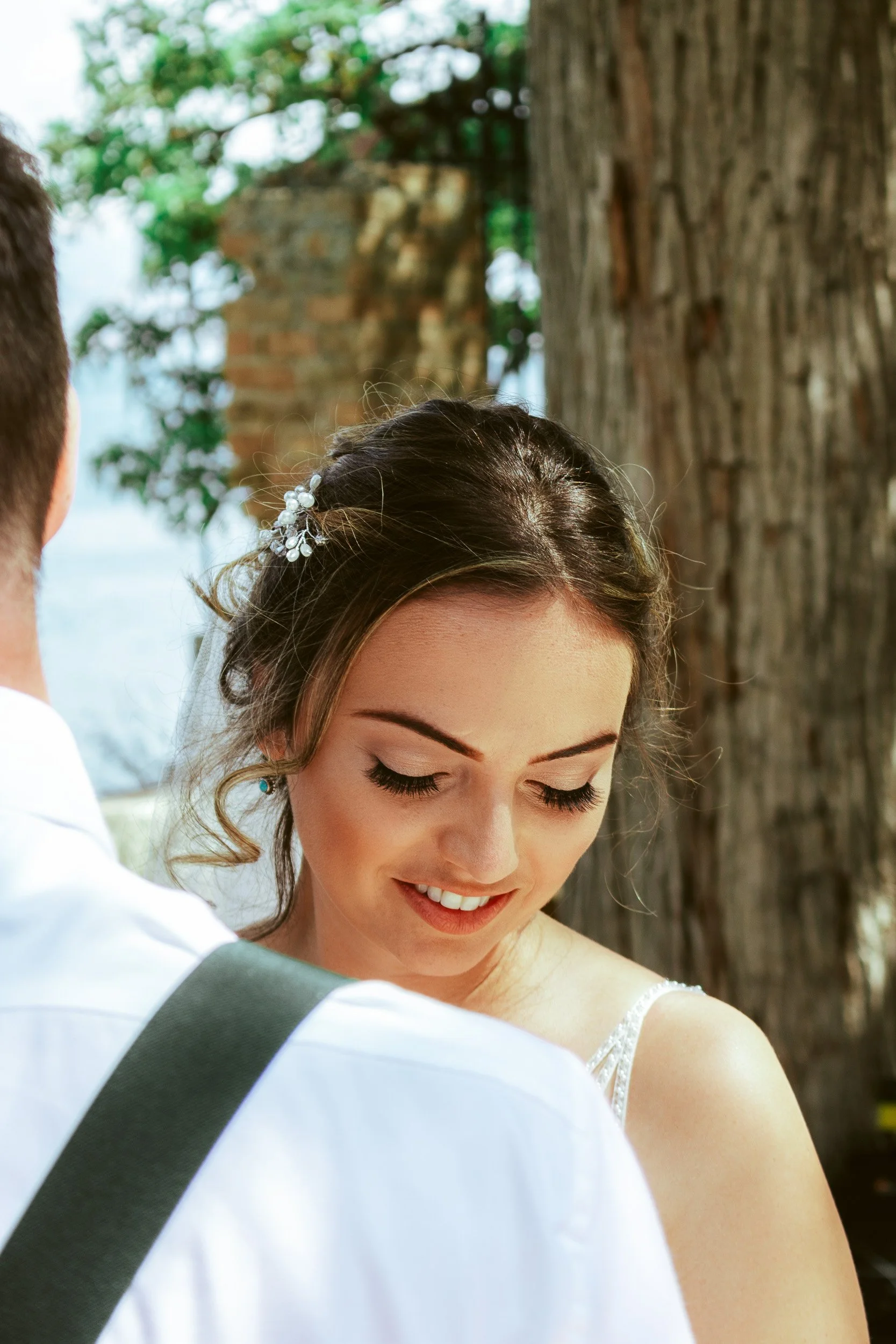 A smiling bride with dark hair and light skin, wearing a white dress and a hair accessory, standing outdoors with trees and a brick structure in the background.