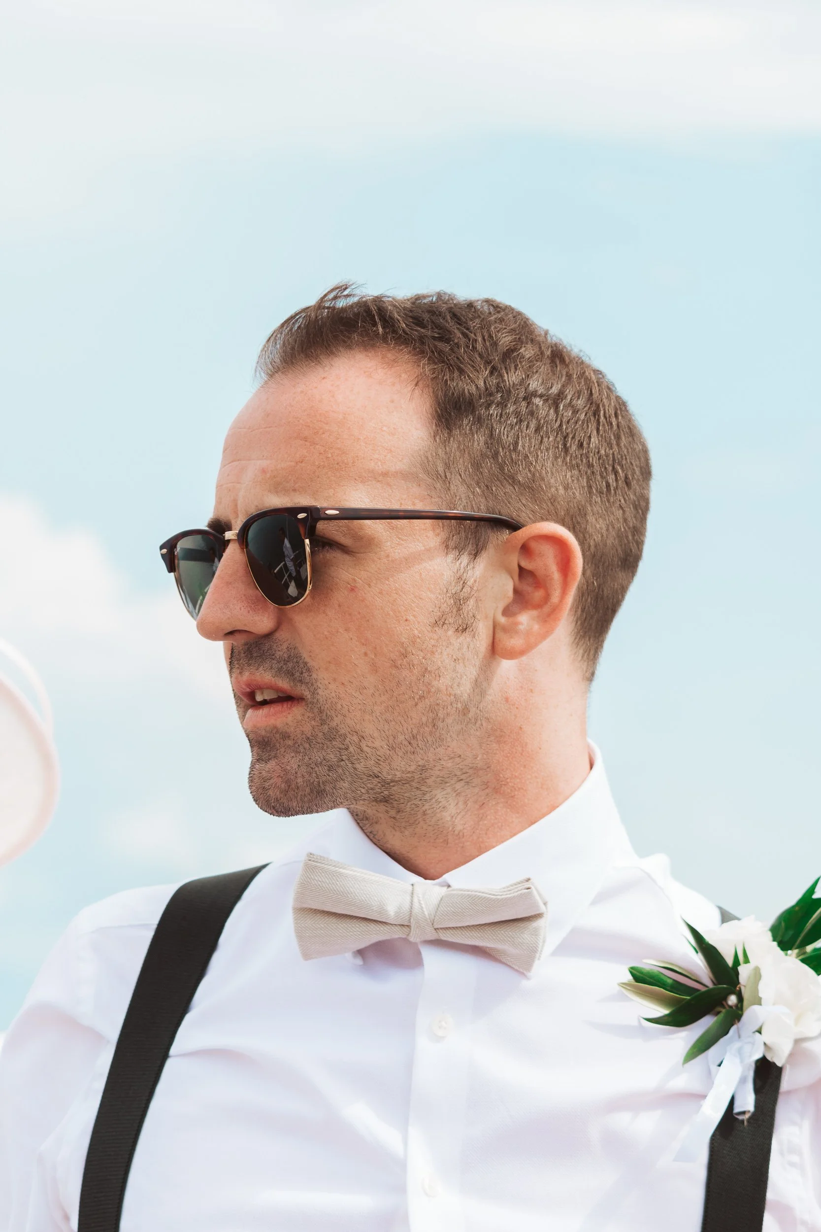 Close-up of a man in a white shirt, beige bow tie, black suspenders, and sunglasses, outdoors against a blue sky.