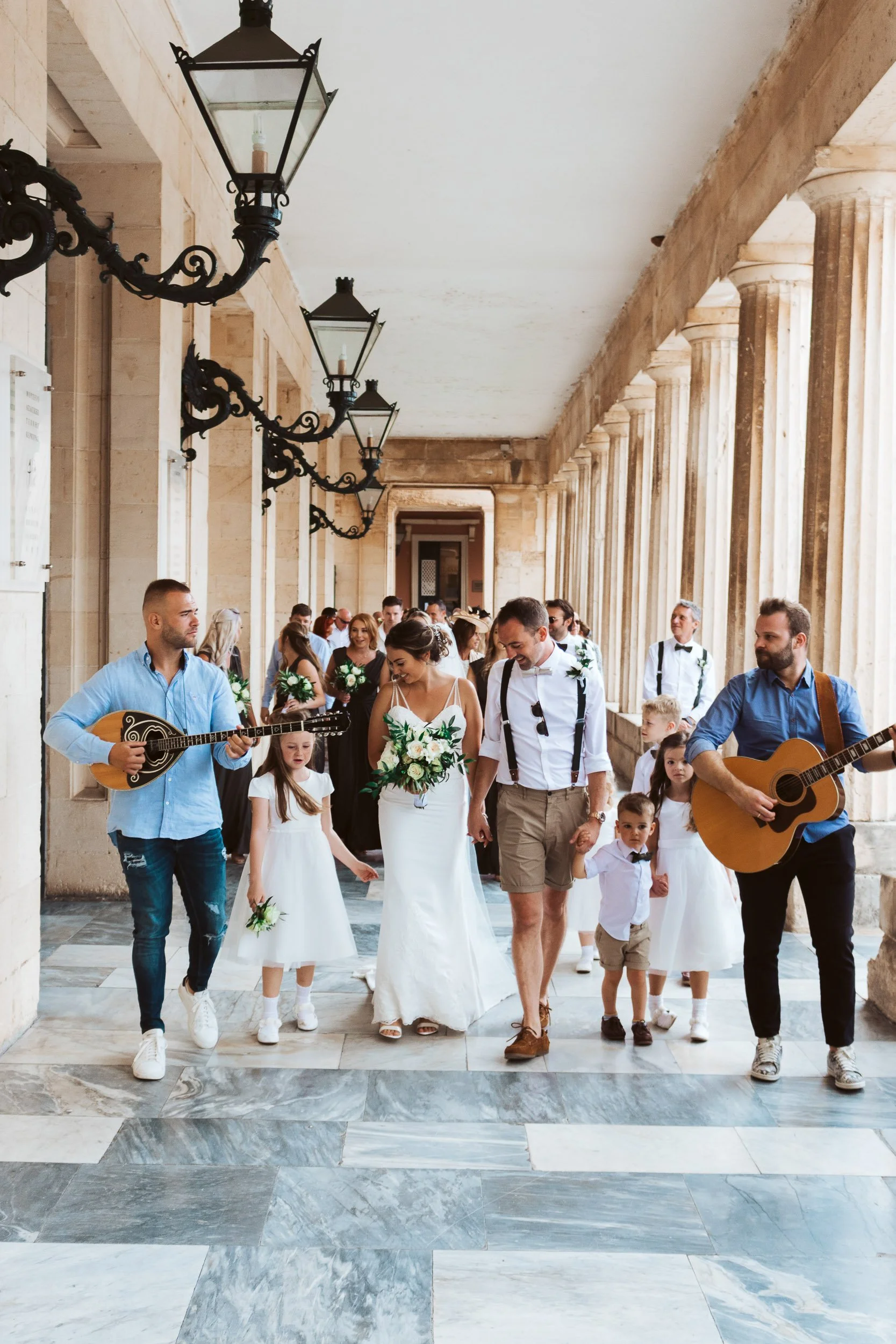 A wedding procession with a bride and groom holding hands, surrounded by children and musicians, walking through a covered outdoor corridor with columned arches and hanging lanterns.