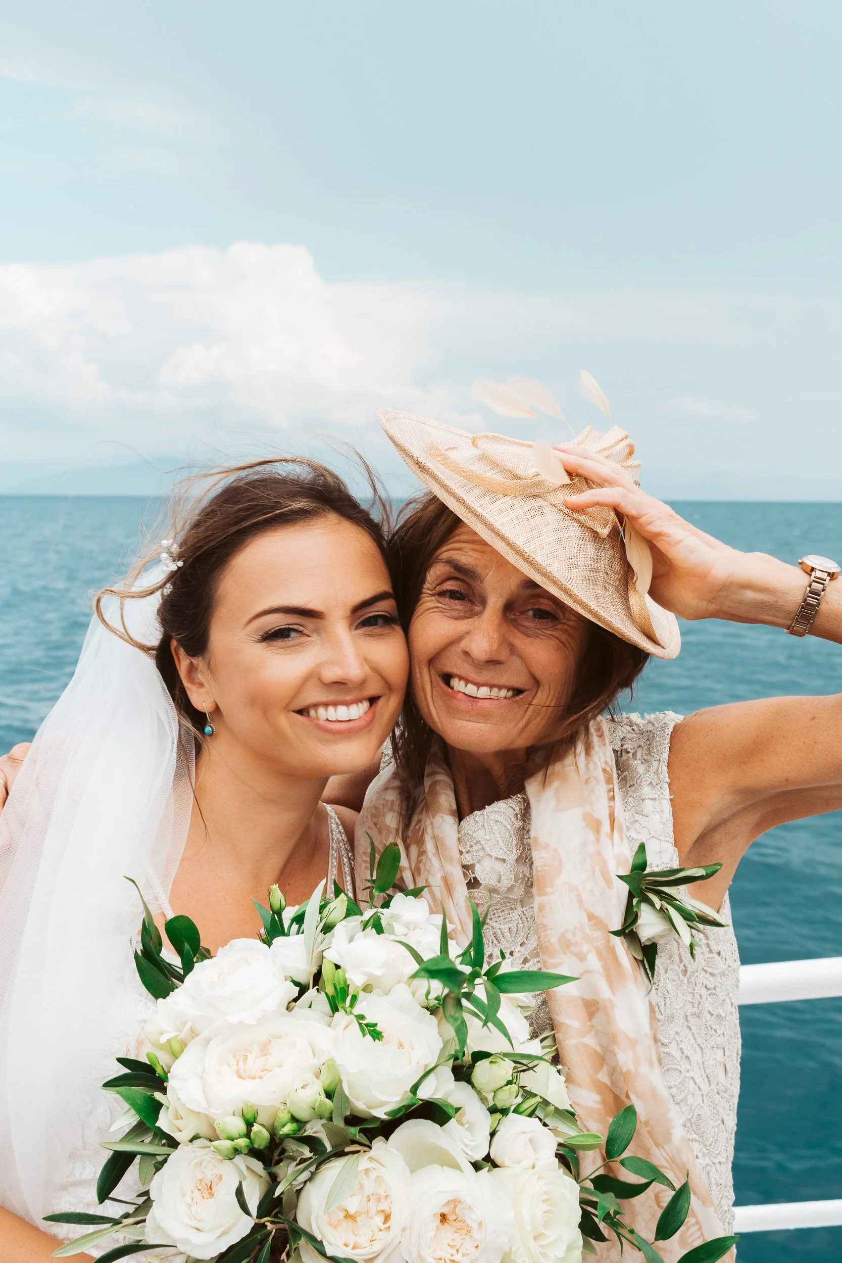 A bride and an elderly woman are taking a selfie on a boat or near water. The bride is holding a bouquet of white flowers, and the elderly woman is wearing a straw hat and a light-colored scarf. Both are smiling, with the water and sky in the backgro