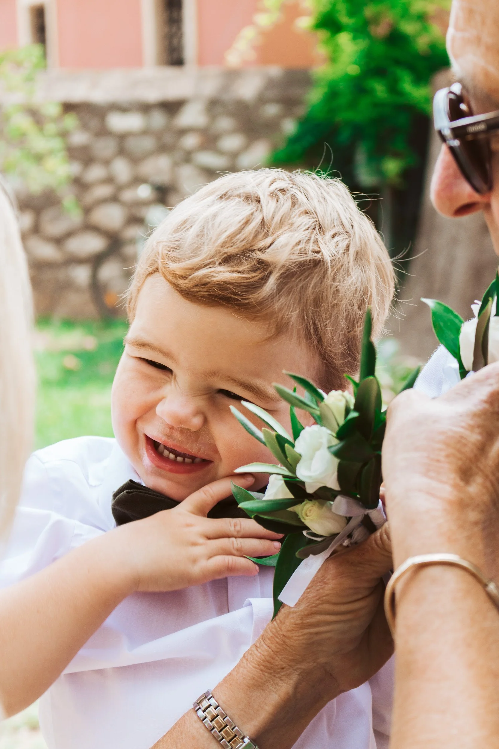 A young boy smiling happily as an older woman gives him a bouquet of white flowers during a celebration outdoors.