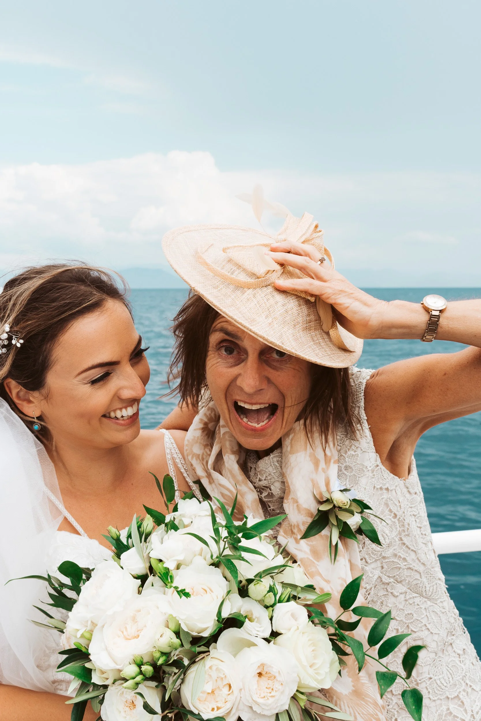 A bride holding a bouquet of white flowers is smiling as an older woman in a hat playfully adjusts it on a boat, with the ocean in the background.