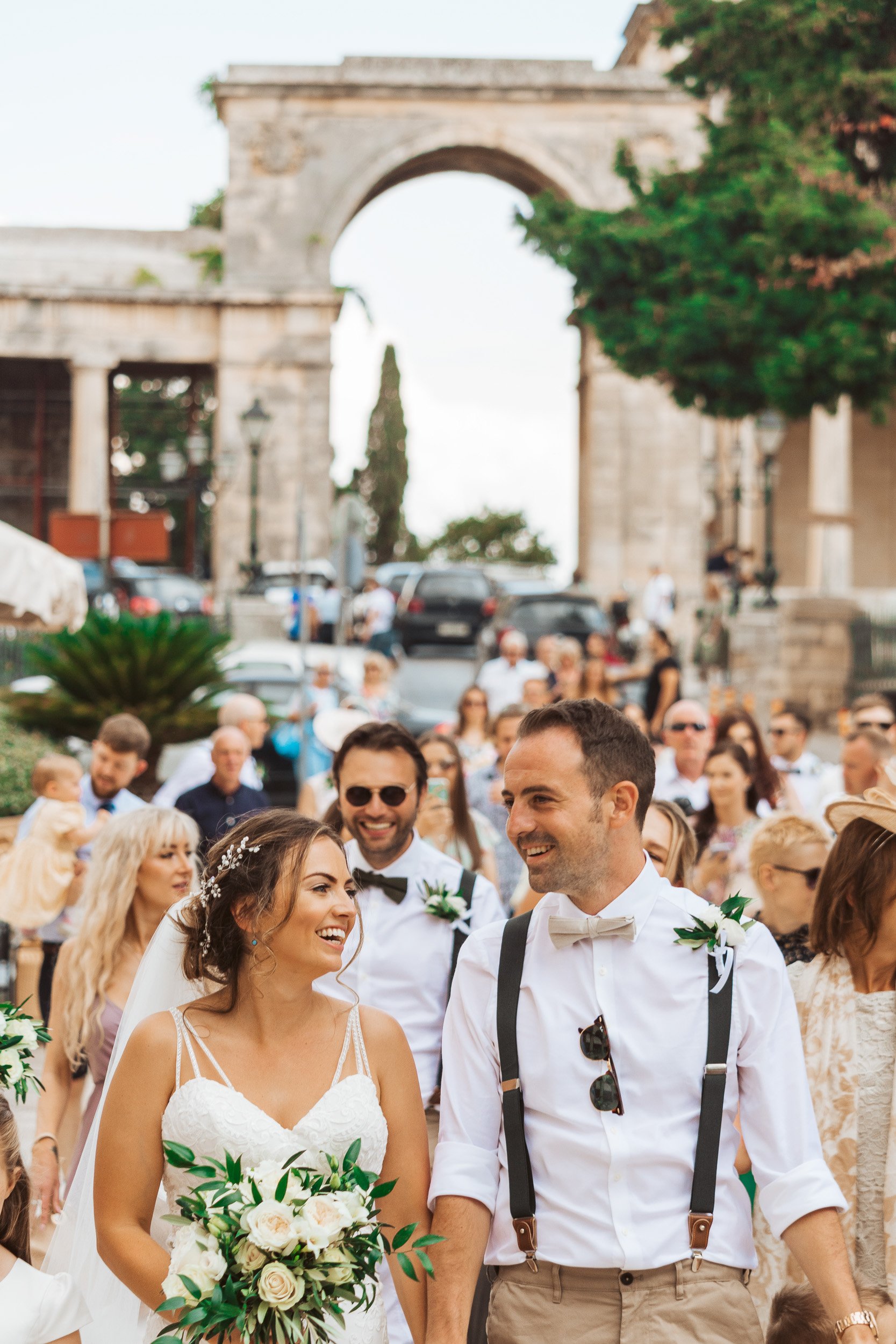 A wedding celebration with a bride in a white dress holding a bouquet and a groom in a white shirt with suspenders, smiling among guests, in front of an ancient stone archway.