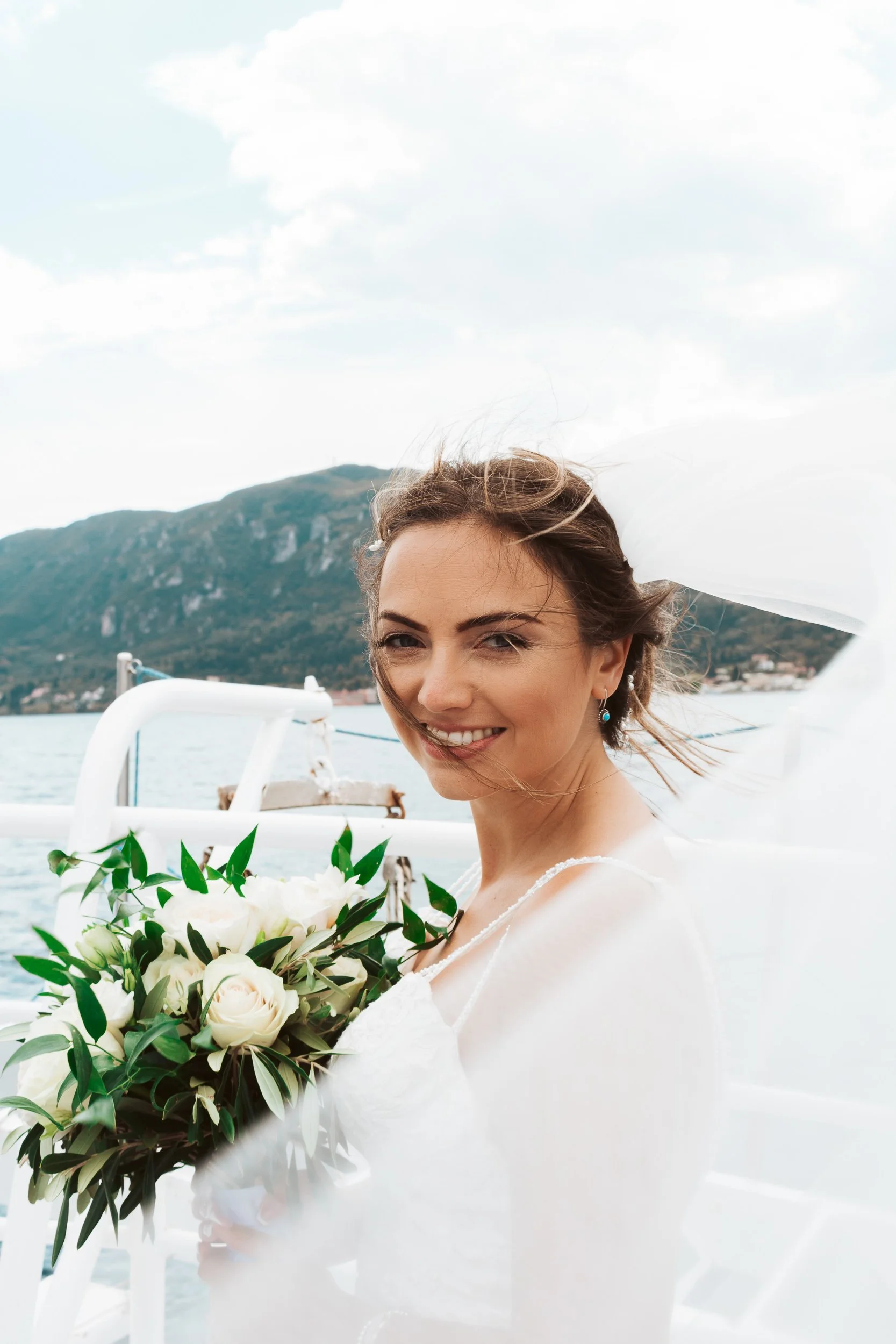 A young woman in a wedding dress holding a bouquet of white roses, smiling on a boat with a mountain and lake in the background.
