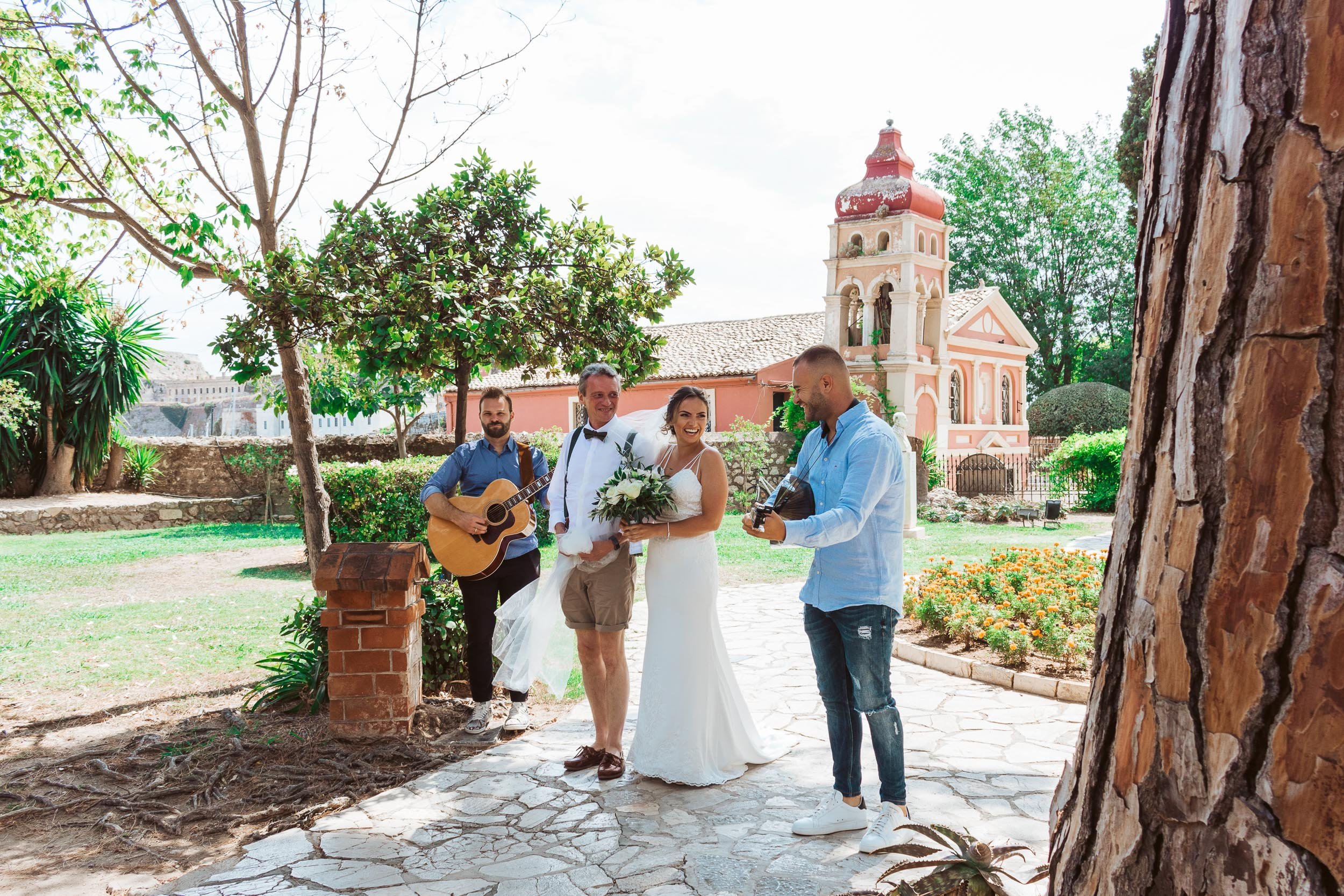 A wedding ceremony outdoors with a bride in a white gown holding a bouquet, a groom in shorts and a bow tie, a man playing guitar, and another man singing, set against a backdrop of a pink chapel and greenery.