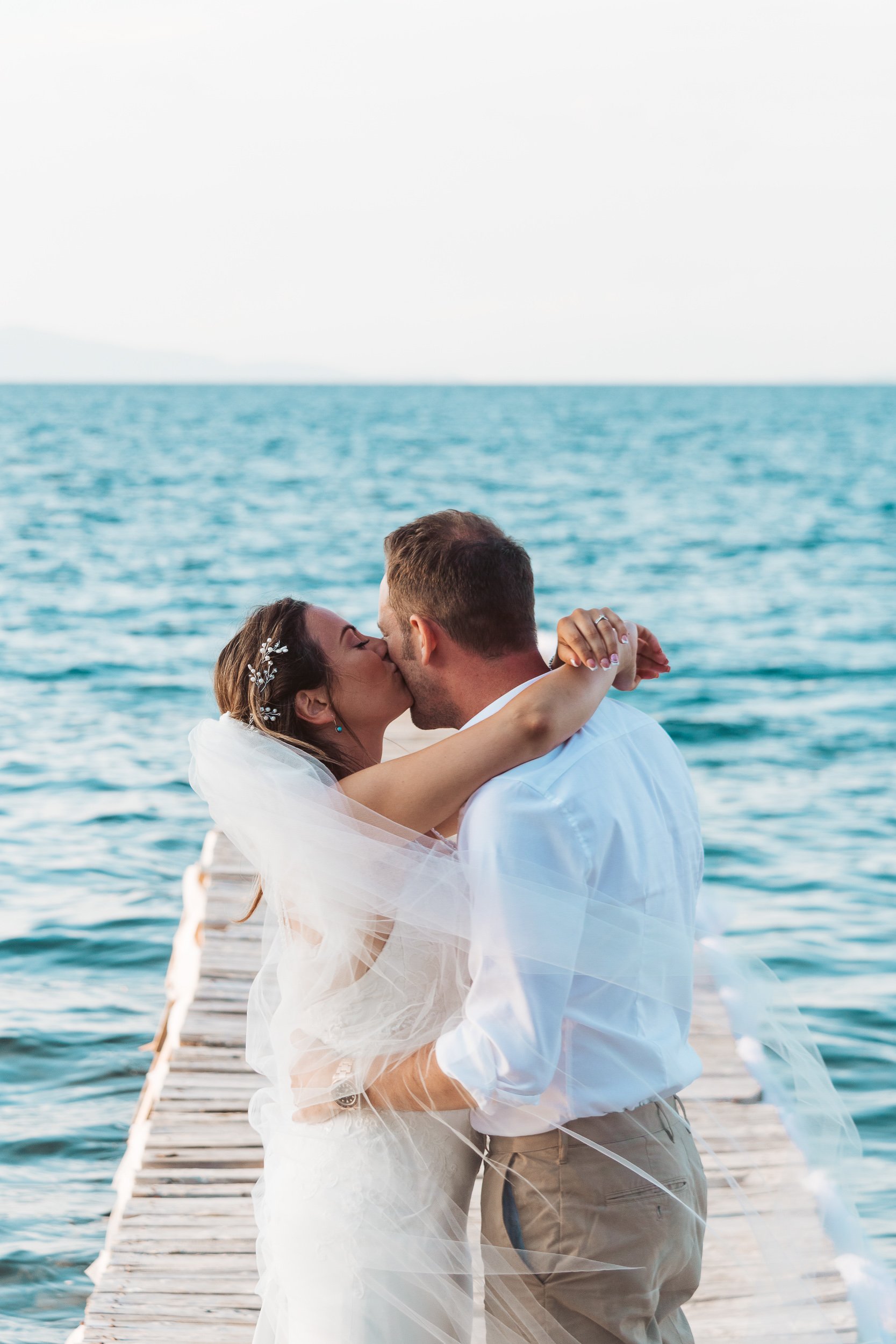 A couple in wedding attire sharing a kiss on a dock by the water during sunset.