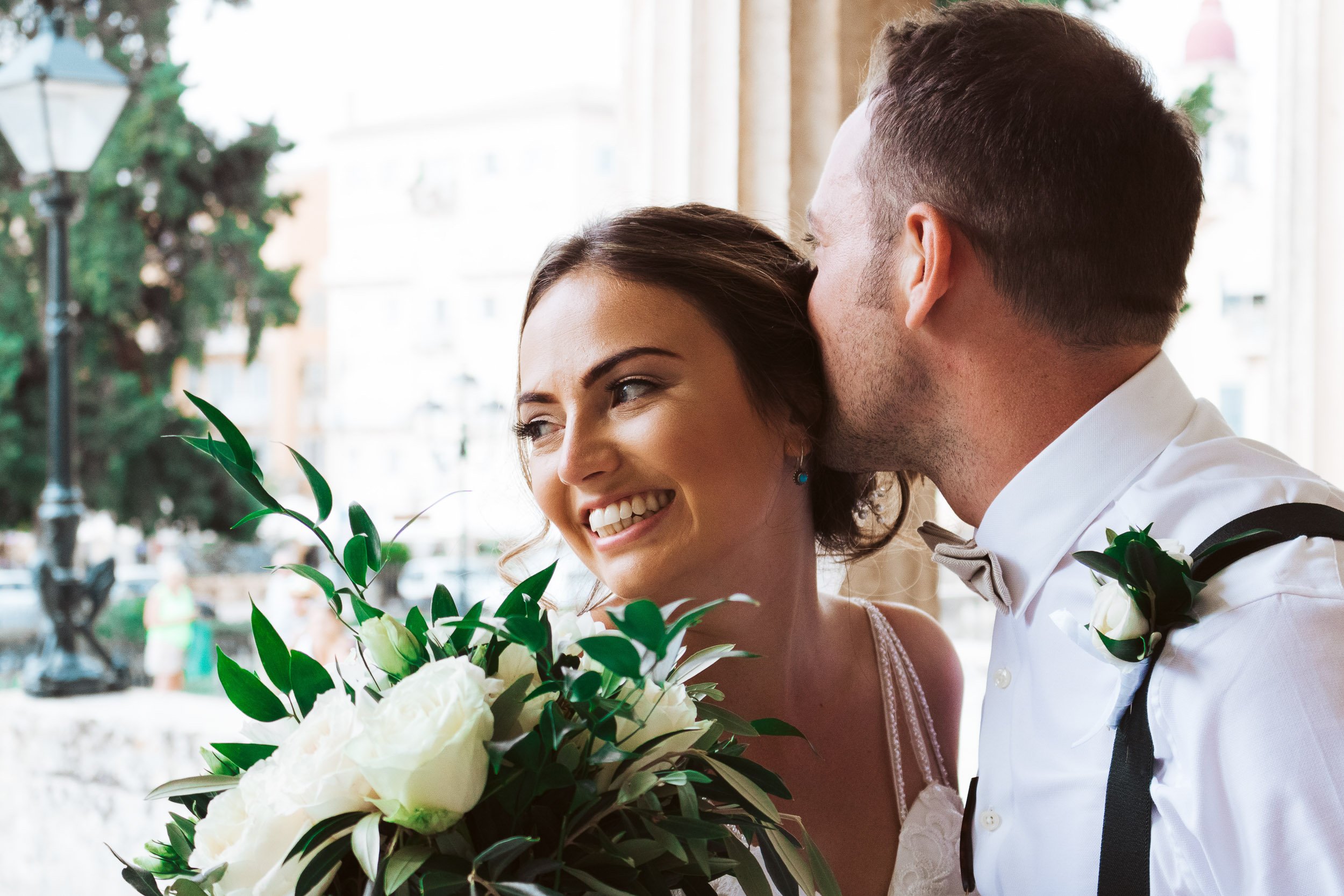 A bride and groom share a joyful moment, with the groom kissing the bride's forehead and the bride smiling, holding a large bouquet of white flowers.
