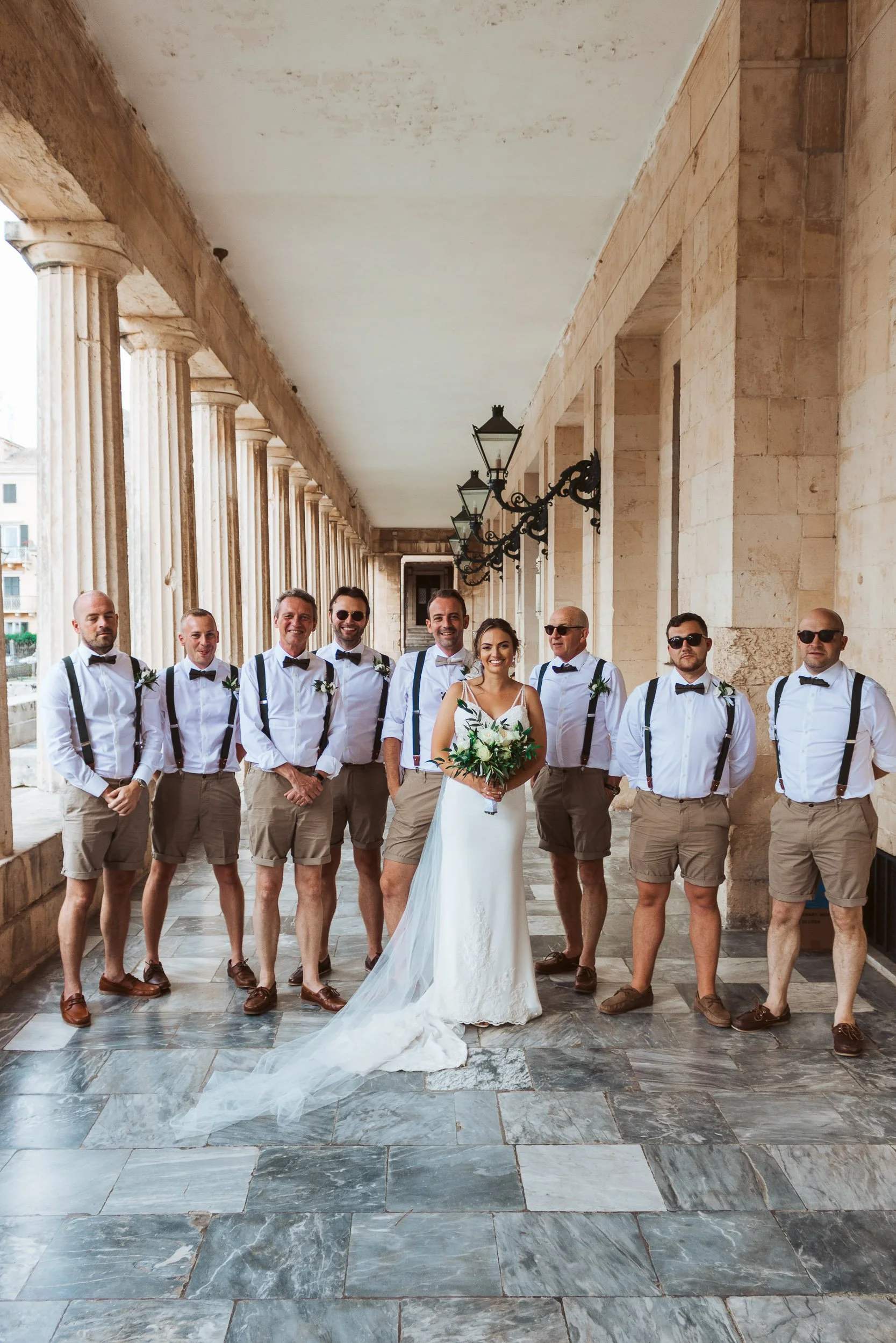 A bride in a white wedding dress holding a bouquet standing with nine men in white shirts, black suspenders, black bow ties, and khaki shorts on a covered stone patio with arches and black wrought iron lanterns.