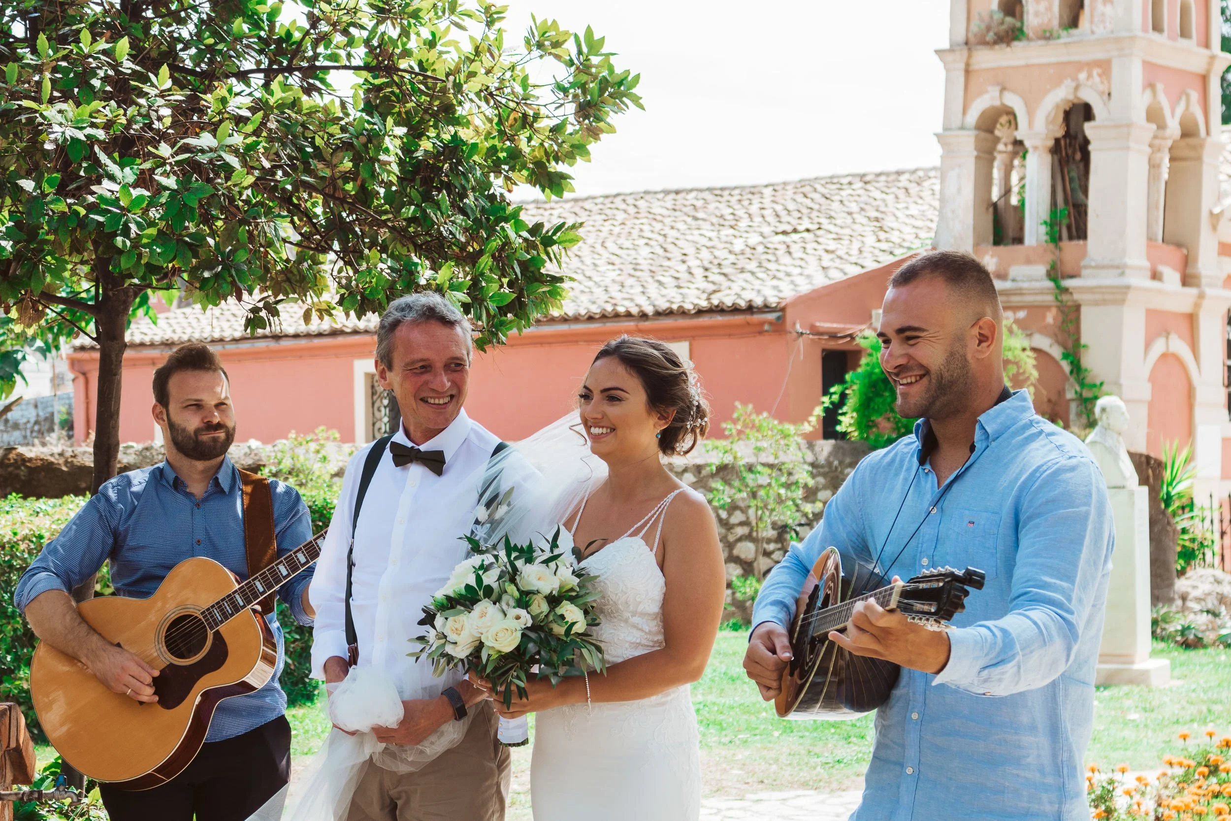 A bride and a groom standing outdoors, smiling, with two musicians playing guitars nearby during a wedding celebration.