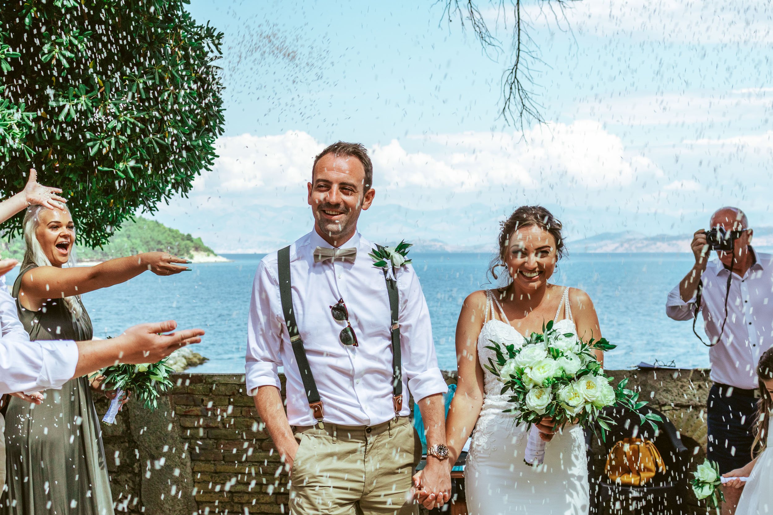 A bride and groom holding hands during their wedding celebration by a body of water, surrounded by friends throwing rice, with a scenic background of water and sky.