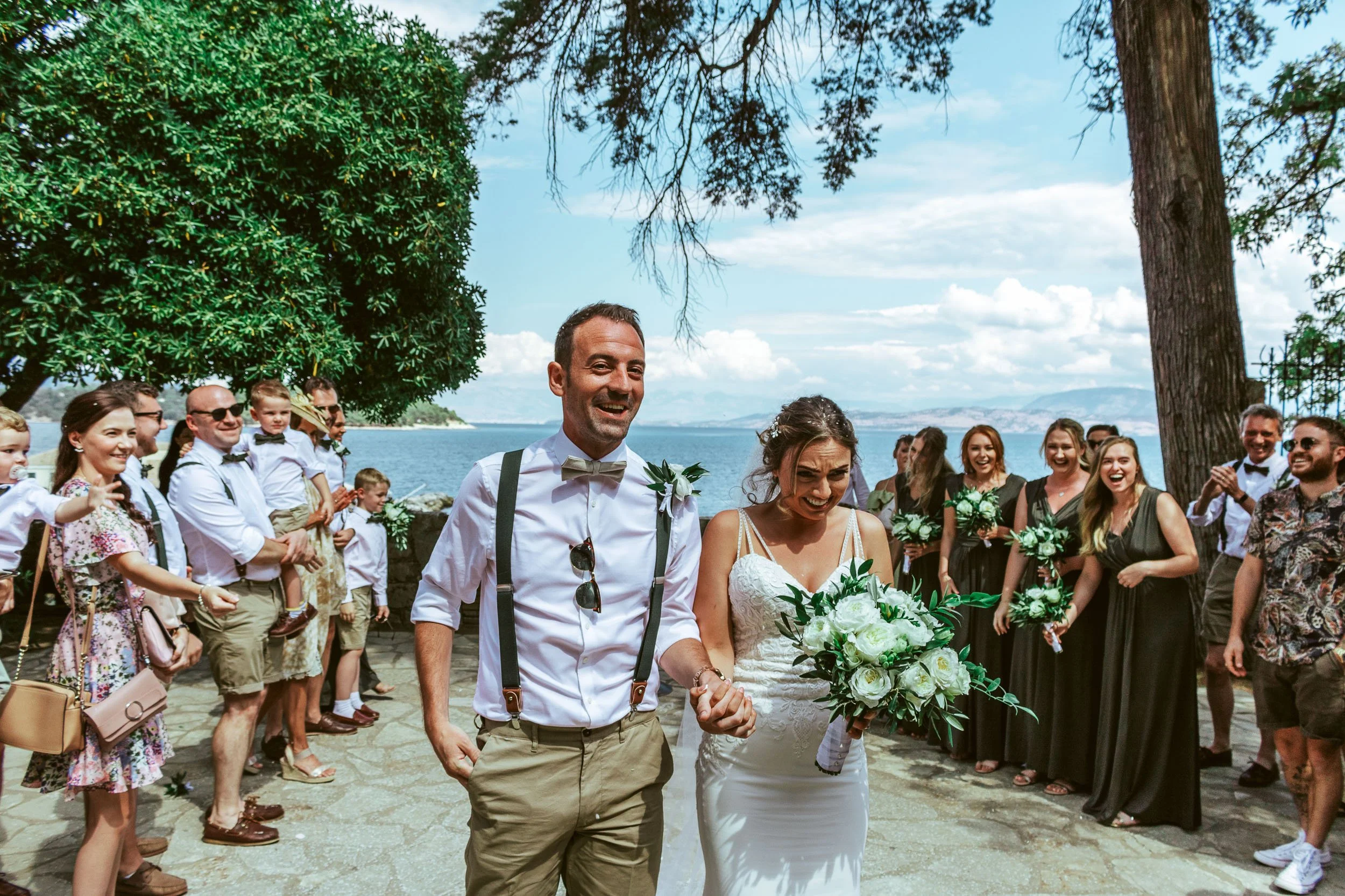 A bride and groom walk hand in hand outdoors near a lake, surrounded by family and friends during a wedding ceremony on a sunny day.