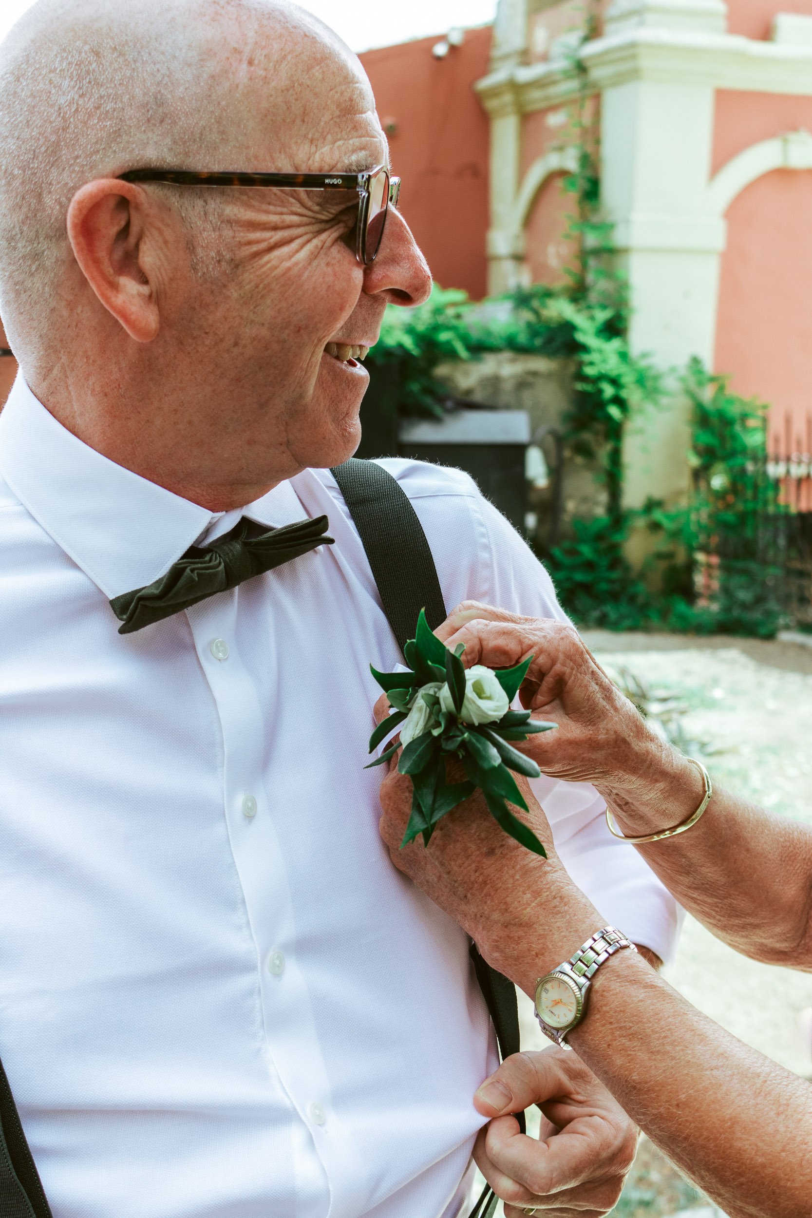 An elderly man dressed in a white shirt and black bowtie, smiling as someone pins a boutonniere with white flowers and green leaves on his shirt. The man is wearing glasses and a wristwatch, with a background of a building and greenery.