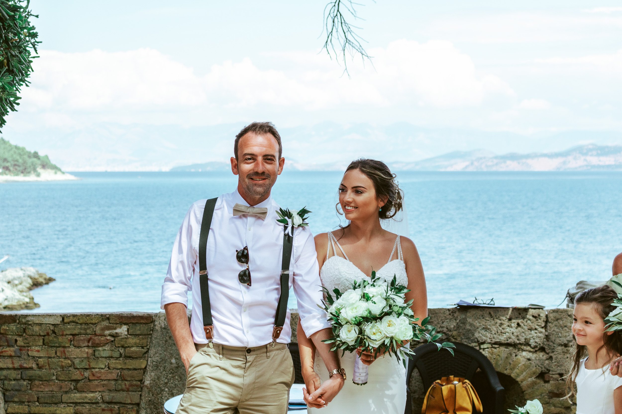 A happy couple at their wedding by a waterfront, with a man in a white shirt, bow tie, and suspenders, and a woman in a white wedding dress holding a bouquet of white roses, with a young girl nearby.