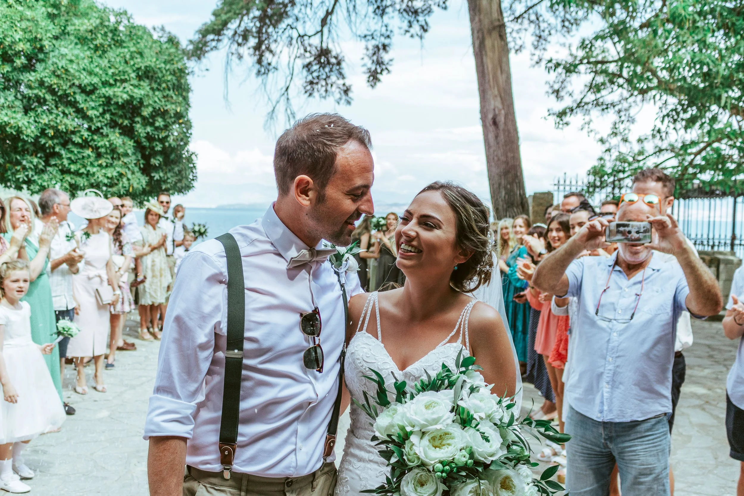 A bride and groom smile at each other during their outdoor wedding, surrounded by friends and family, with a scenic waterfront in the background.
