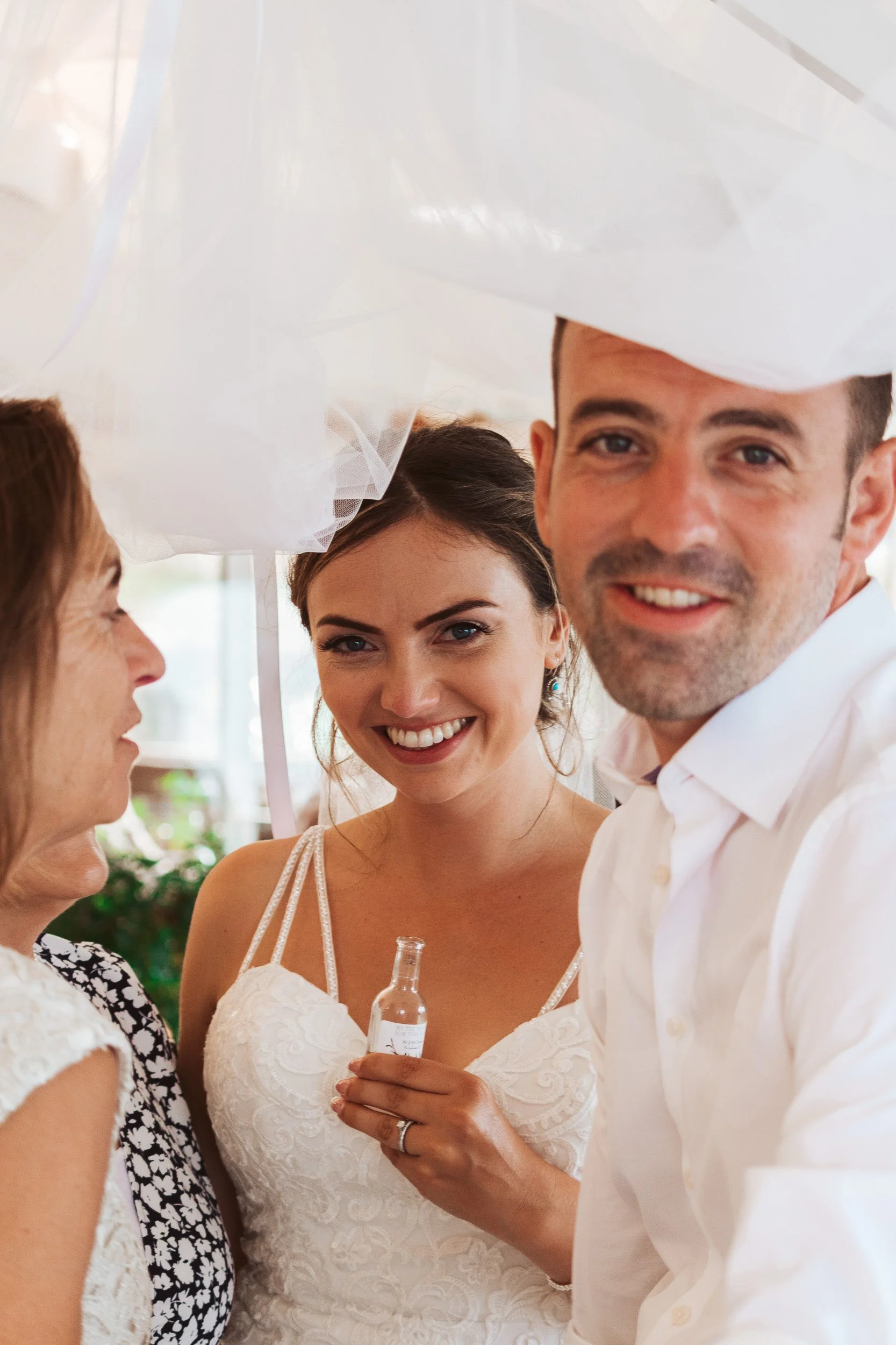 Smiling woman in wedding dress holding small bottle, flanked by a man in white shirt and an elderly woman, at a celebration under white decor.