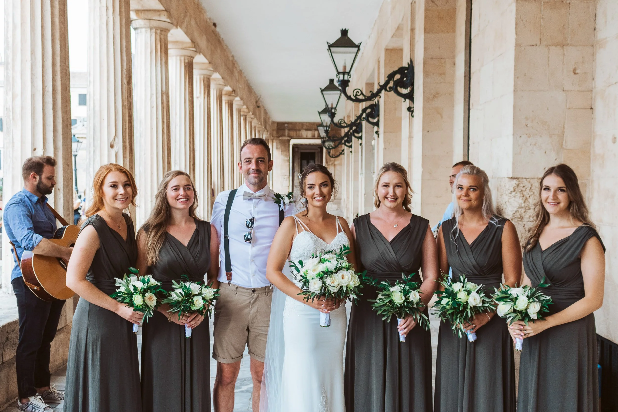 A group of seven people, including a bride and groom, standing in a covered outdoor corridor with stone columns and lantern-style lights, smiling at the camera. The bride is holding a bouquet of white flowers, and four women beside her are holding ma