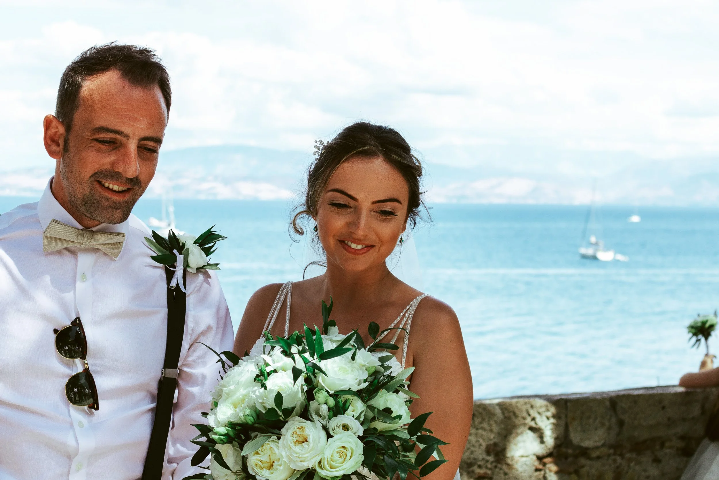 A bride and groom at a wedding, standing outdoors near a body of water with sailboats, holding a bouquet of white roses and greenery, smiling.