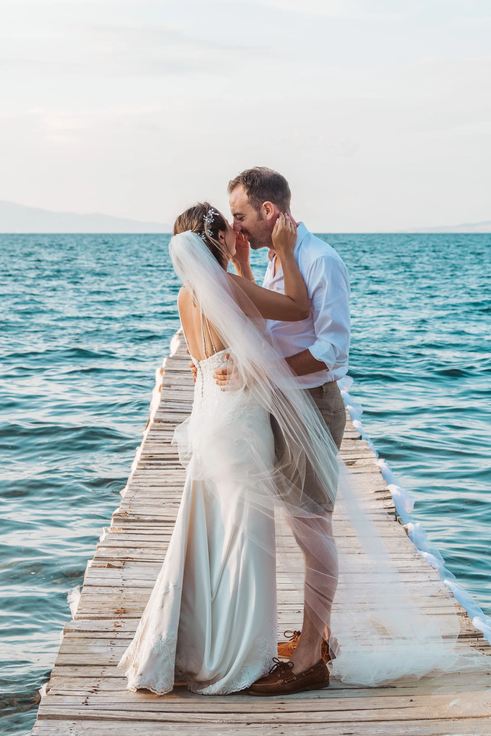 A bride and groom embrace on a wooden dock extending into the water, with mountains in the distance and the sky overhead.