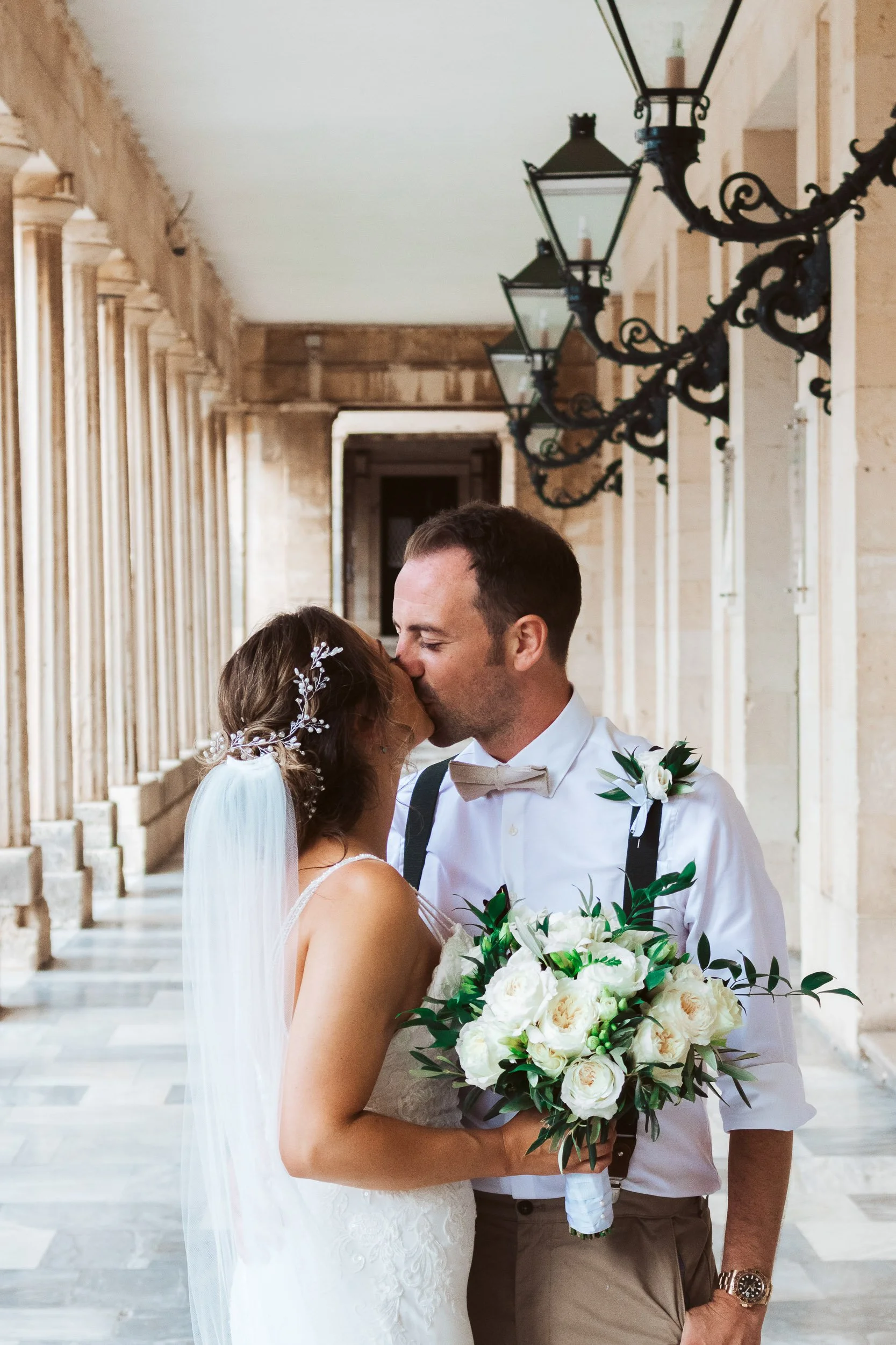 A newlywed couple sharing a kiss in a covered outdoor corridor, with vintage-style lanterns hanging above and stone walls on either side. The bride is holding a bouquet of white roses and greenery.