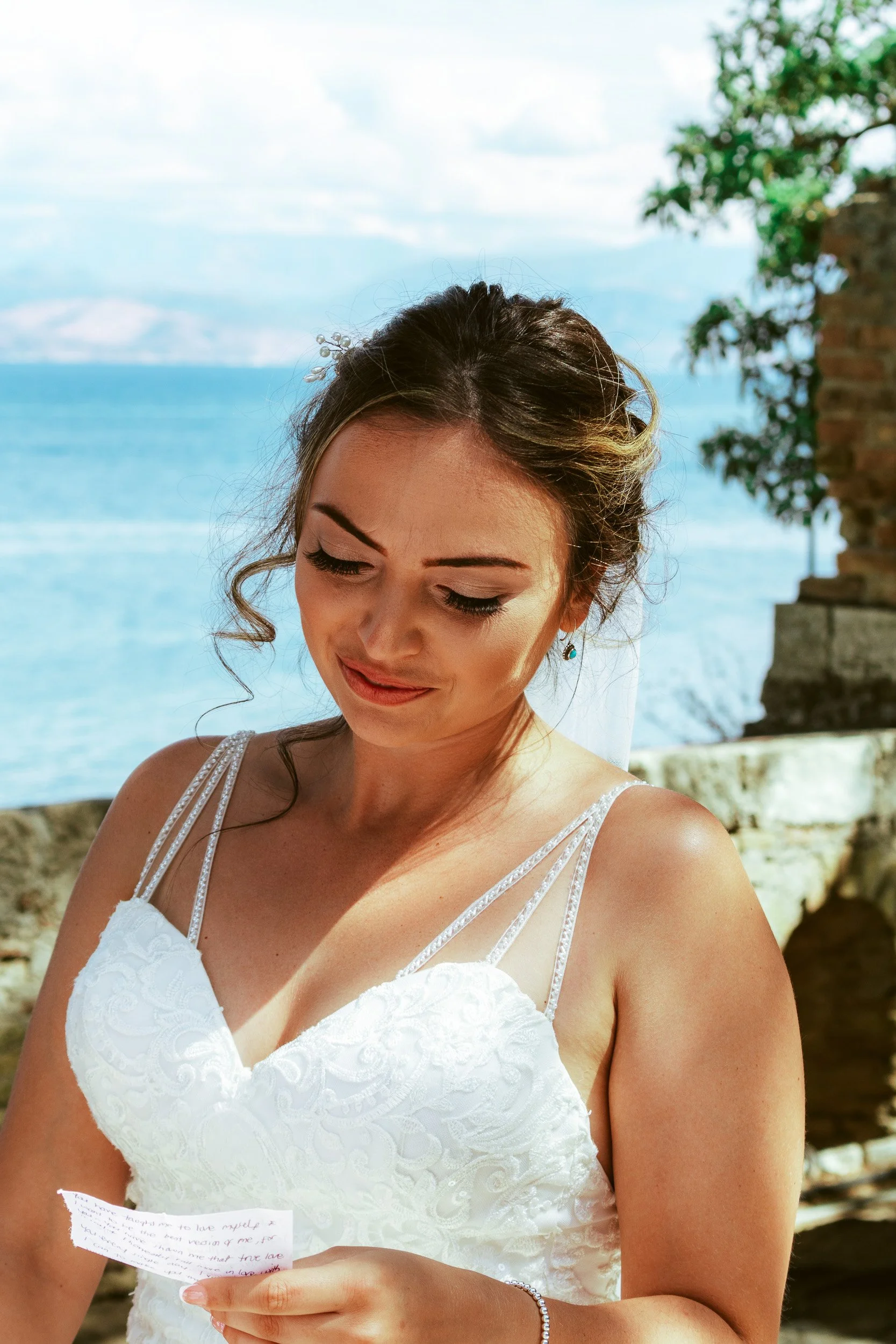 A bride in a white wedding dress holding a handwritten note, standing outdoors by the water with a sunset or early evening sky in the background.