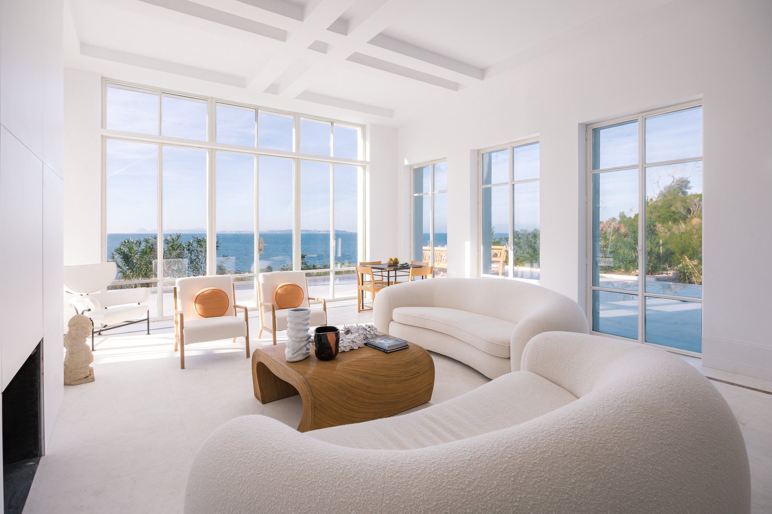 Bright living room with large windows showing ocean view, white sofas, wooden coffee table, and chairs near the windows.