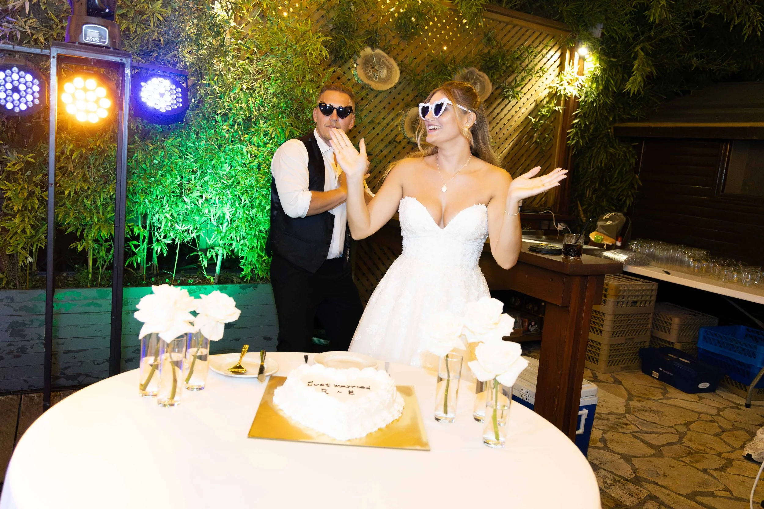 A newlywed couple celebrating at their wedding reception. The bride is in a strapless white wedding dress and wearing heart-shaped sunglasses, smiling and raising her hands. The groom, wearing dark sunglasses, is standing behind her, dressed in a ves
