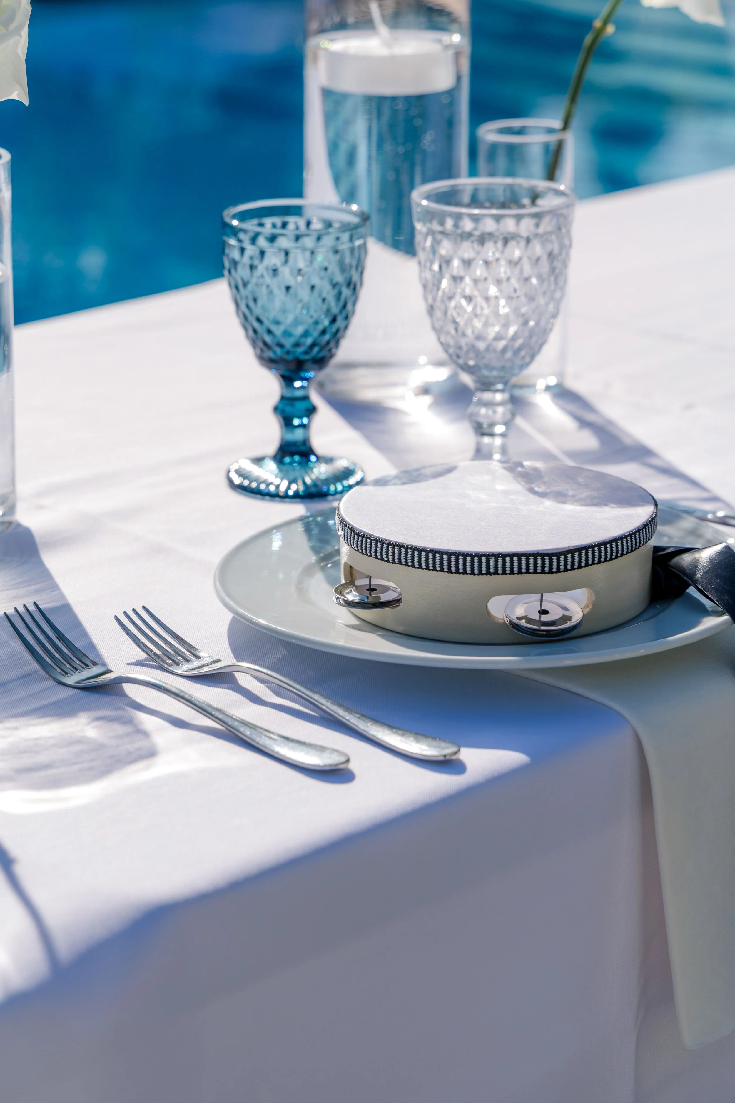 Elegant table setting with white tablecloth, silver forks, blue and clear cut-glass wine glasses, a glass pitcher of water, and a white cushion in a silver case, possibly a ring holder, with a blue-striped ribbon around the edge, set outdoors near a 