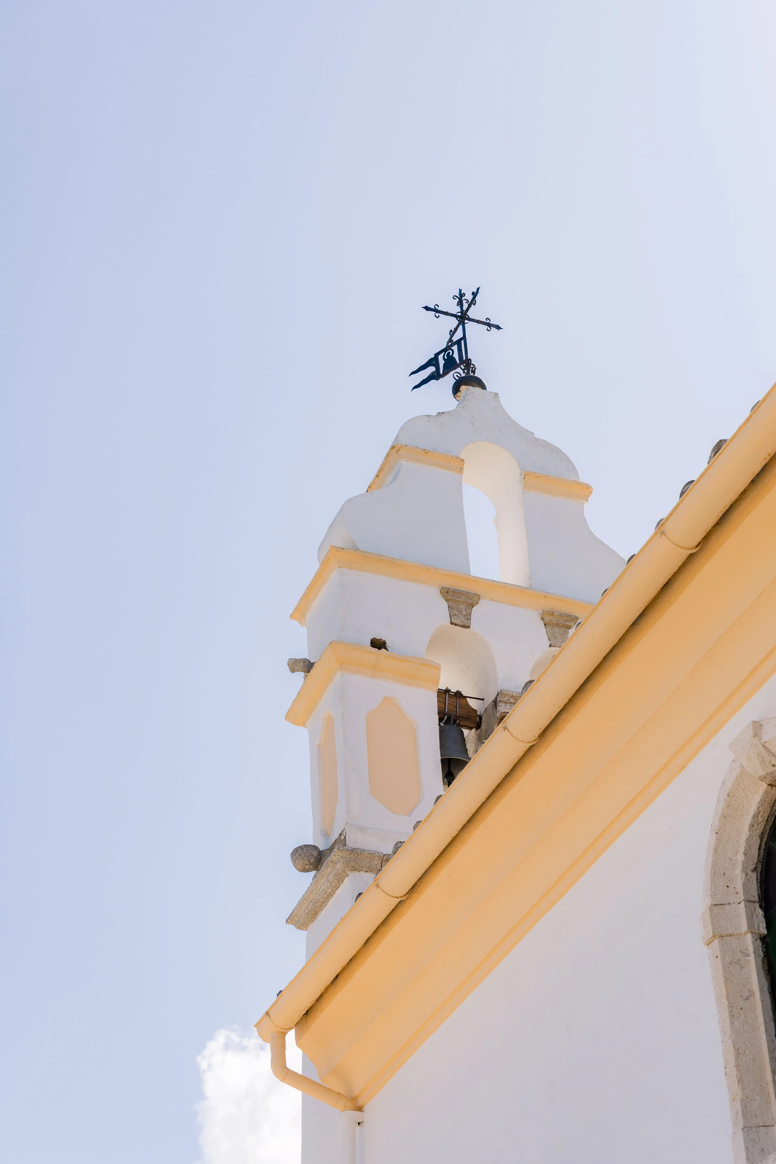 A church steeple with a black weather vane on top against a light blue sky, yellow trim along the edge of the roof, and a small bell visible.
