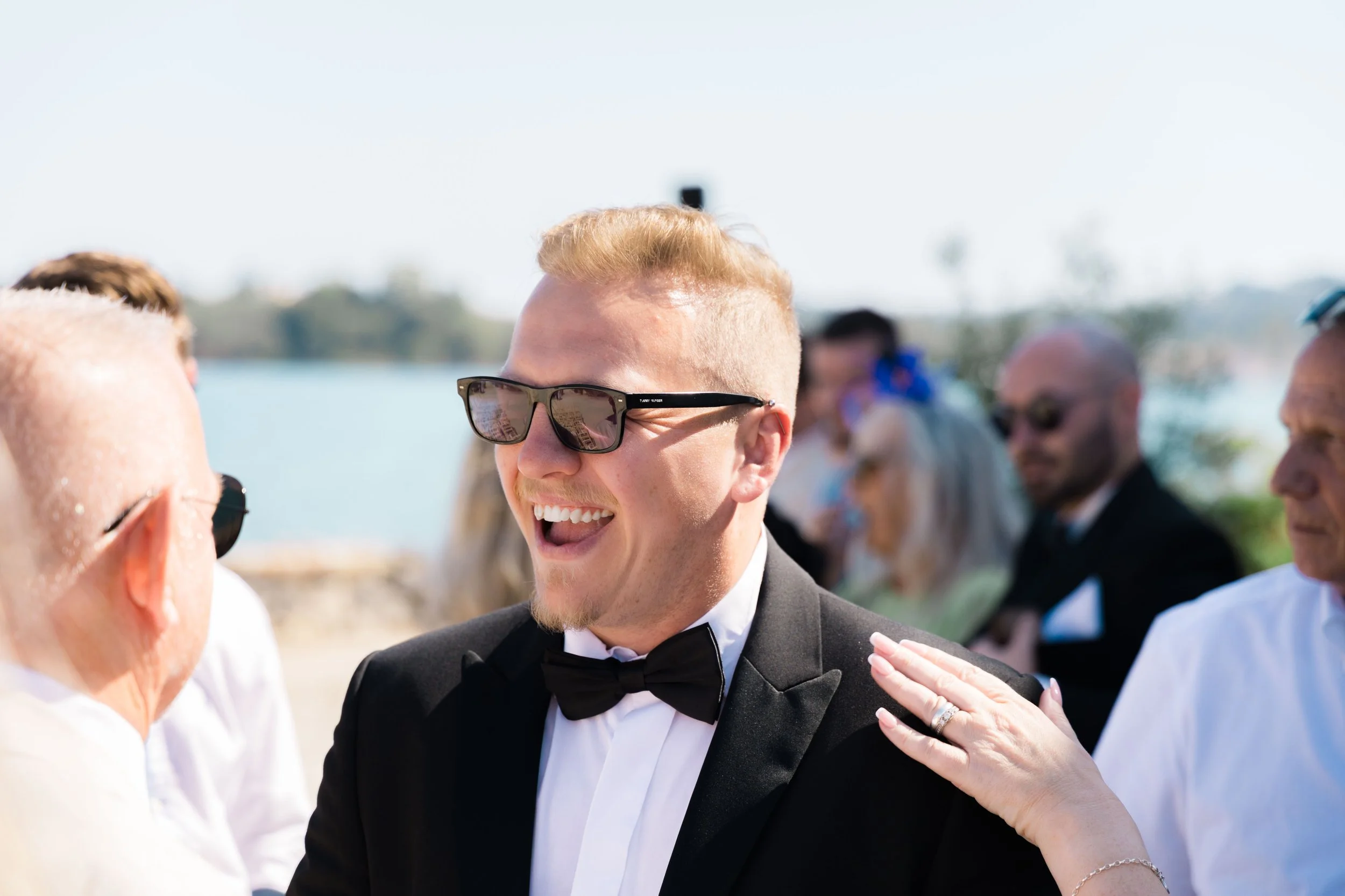 A smiling man in a tuxedo and sunglasses at an outdoor wedding, talking to a woman with her hand on his shoulder. There are other guests in the background by the water.