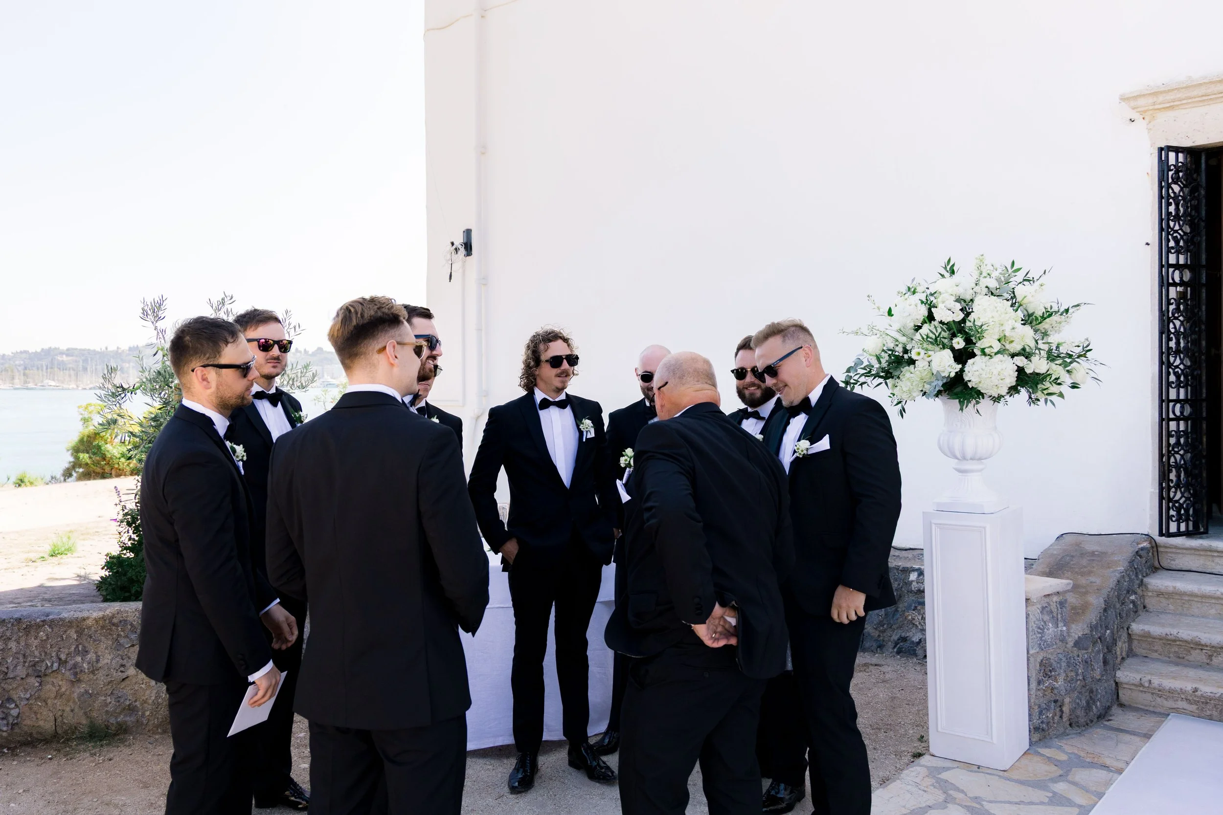 Group of men in black tuxedos and sunglasses standing outdoors in a ceremony or event setting, with a large white floral arrangement on a pedestal nearby.
