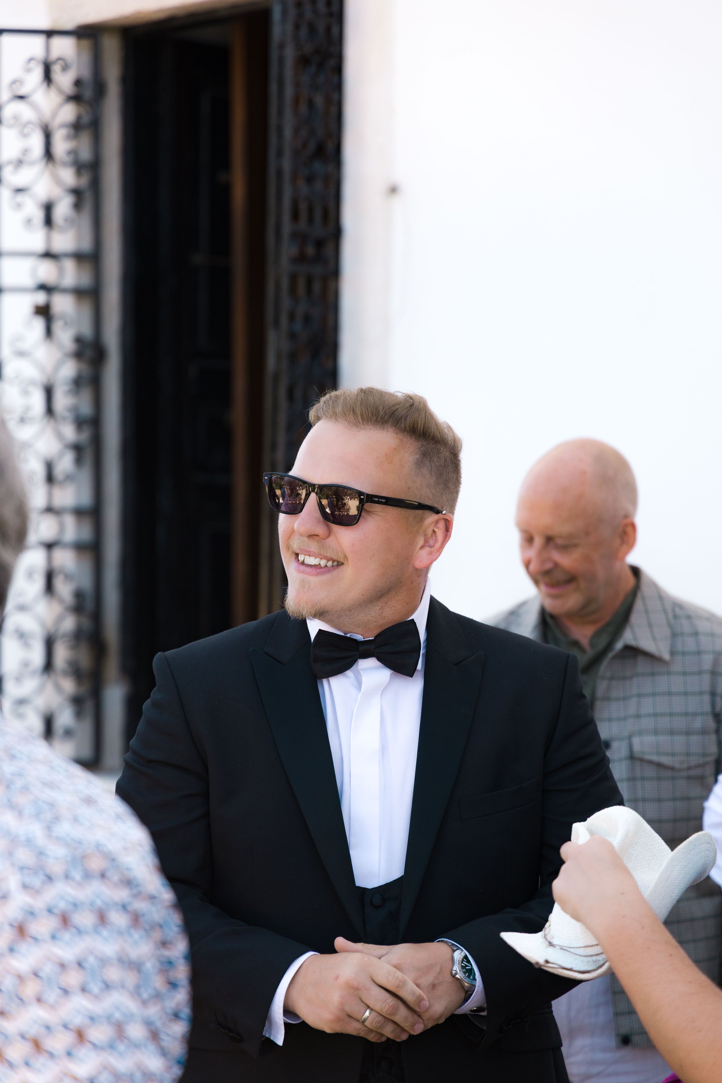 A young man in a black tuxedo with a bow tie and sunglasses smiling, standing outdoors near a group of people, with an older man in a checkered shirt in the background.