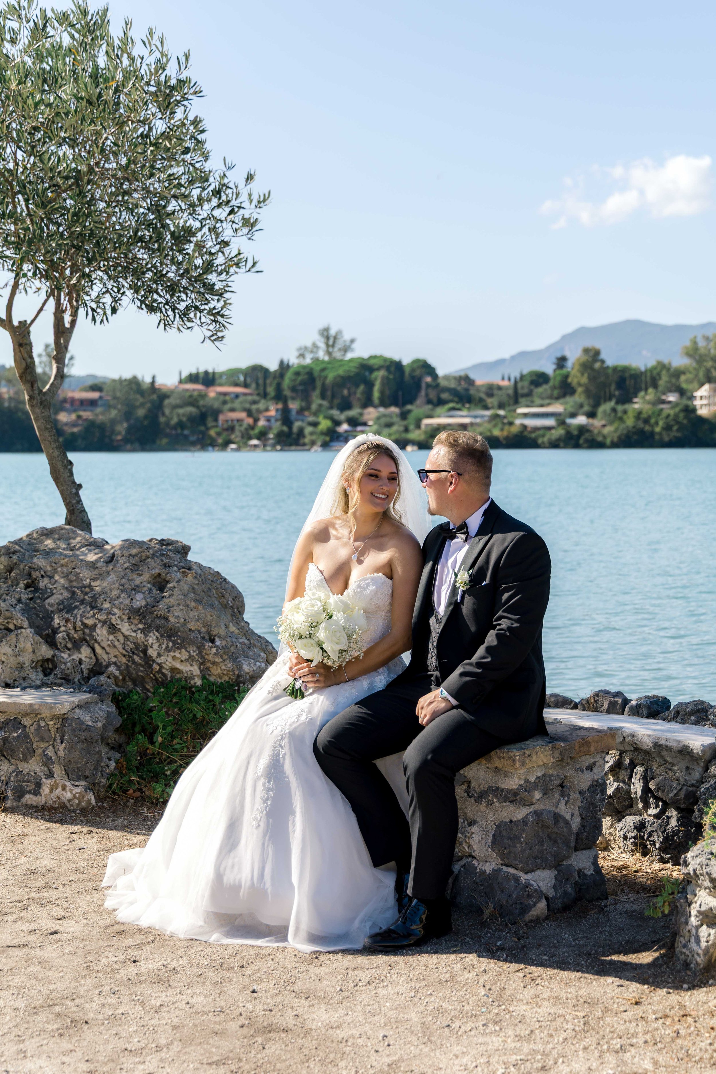 A bride and groom sitting on a stone bench near a lake with trees and hills in the background.