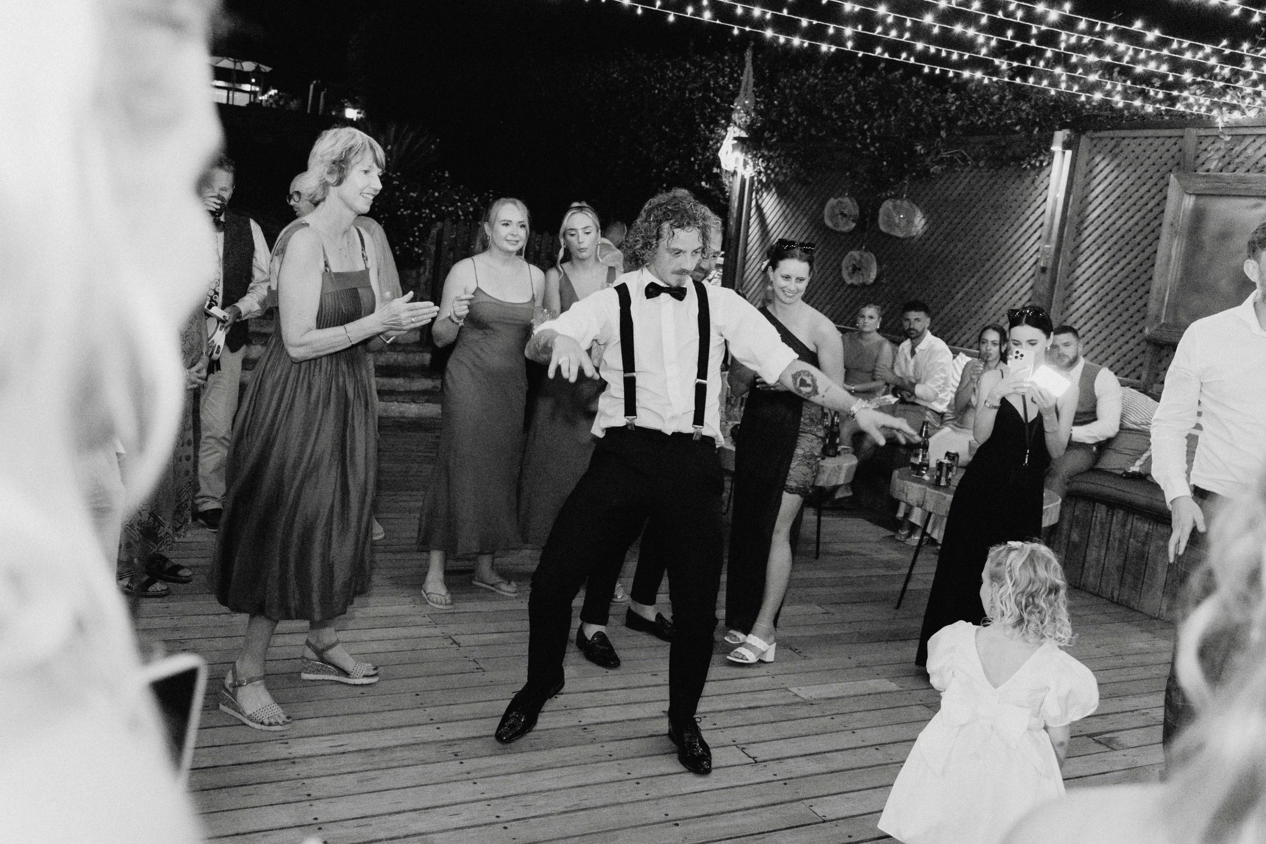A group of people dancing and watching at a party or celebration outdoors at night, decorated with string lights.