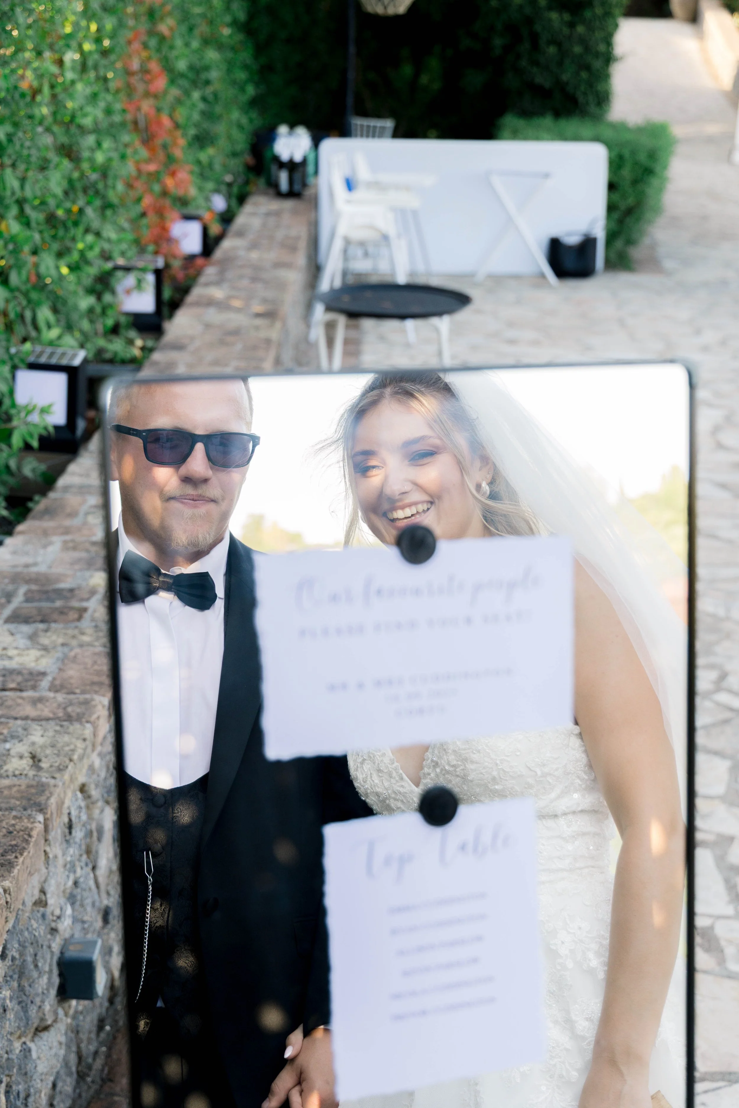 A bride and groom taking a photo through a mirror with wedding signs attached to it, outdoors on a stone patio.