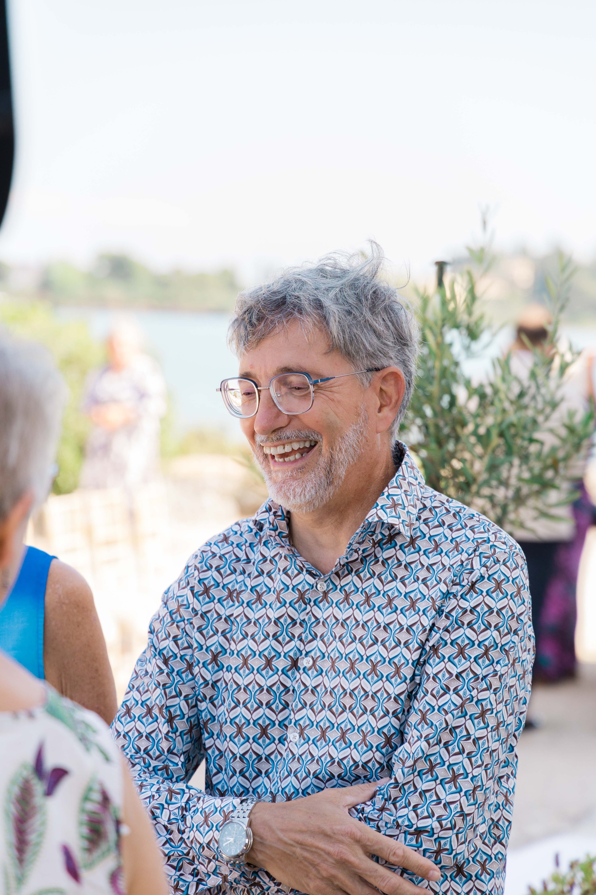 A smiling older man with glasses and gray hair, wearing a patterned shirt, talking at an outdoor social event near a body of water.