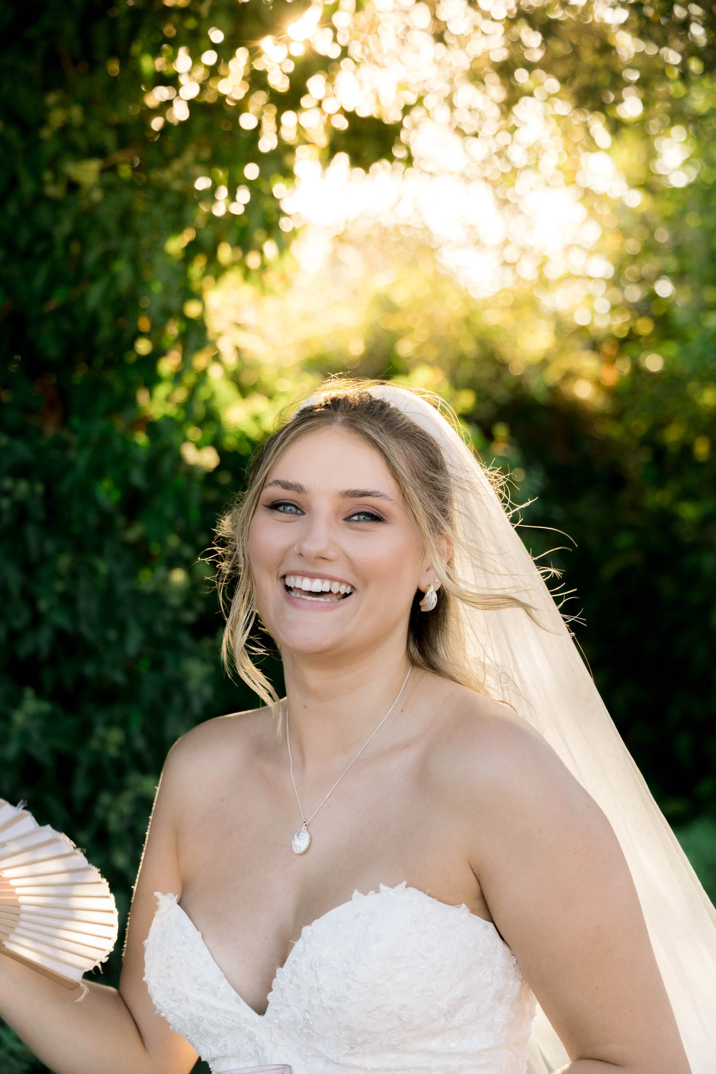 A smiling bride in a strapless white wedding dress, with a veil and jewelry, outdoors during sunset.