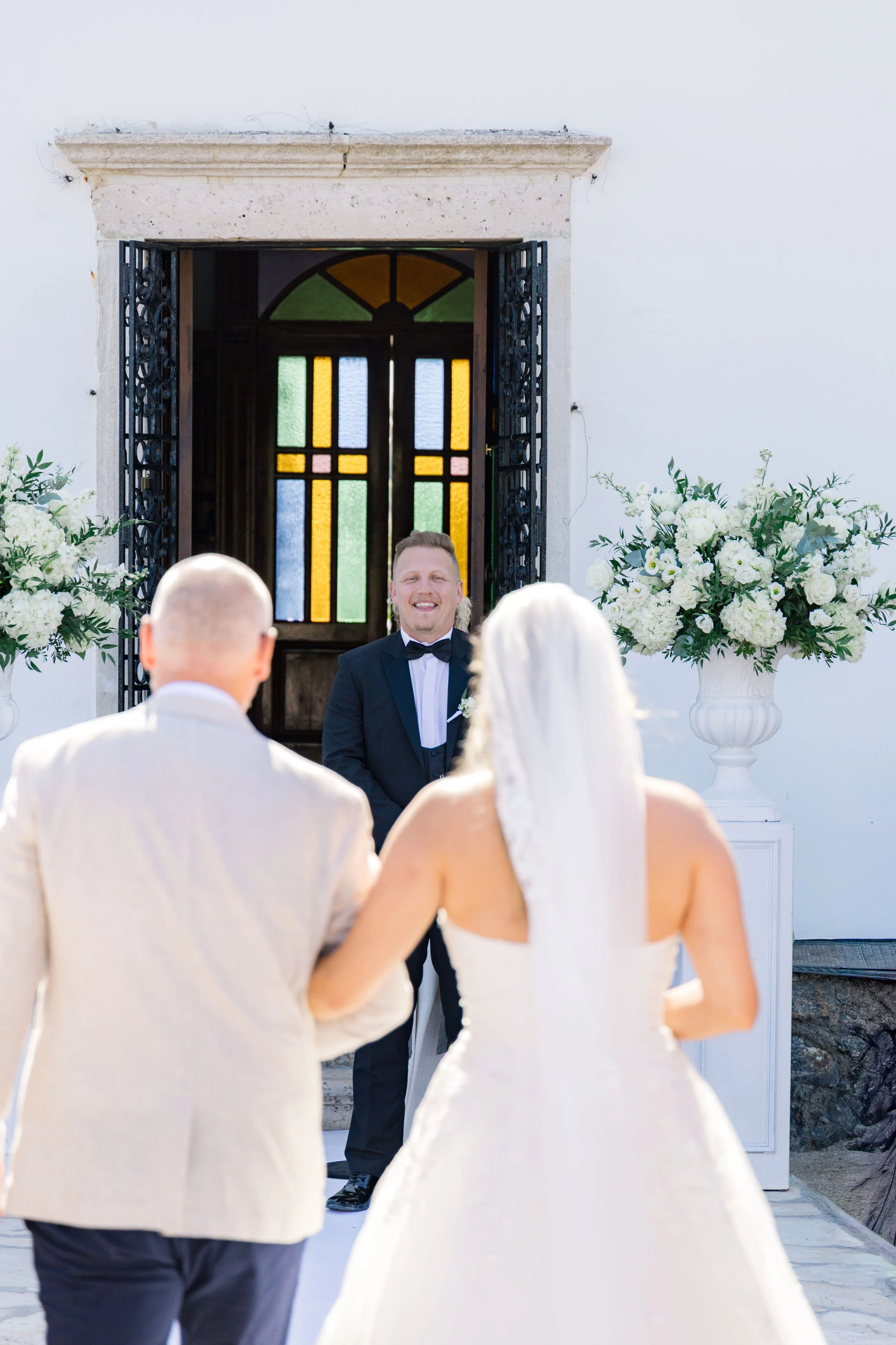 A joyful groom in a black tuxedo stands at the altar during a wedding ceremony, with a bride in a white wedding dress and veil holding his arm, facing an officiant outside a church with white walls, stained glass door, and large white floral arrangem