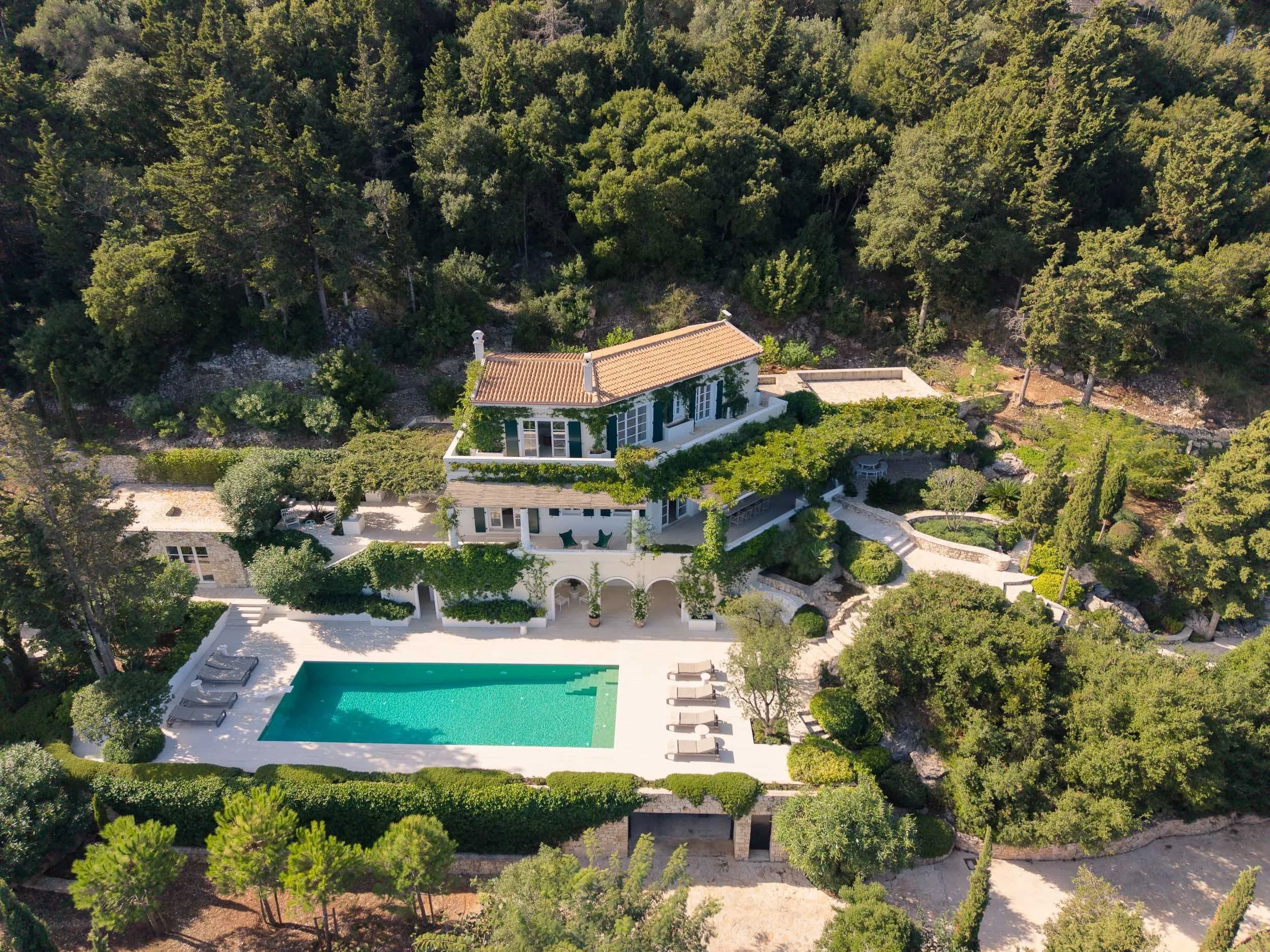 A large white house with multiple levels, terraced gardens, and a swimming pool in a lush, wooded area.